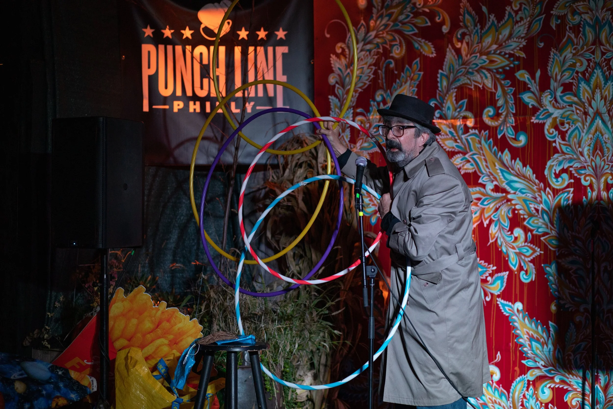 A man with glasses, a beard, and a hat, wearing a trench coat, stands on a stage with a microphone, spinning colorful hula hoops. The background displays a sign that reads "Punch Line Philly" and a red, decorative curtain.
