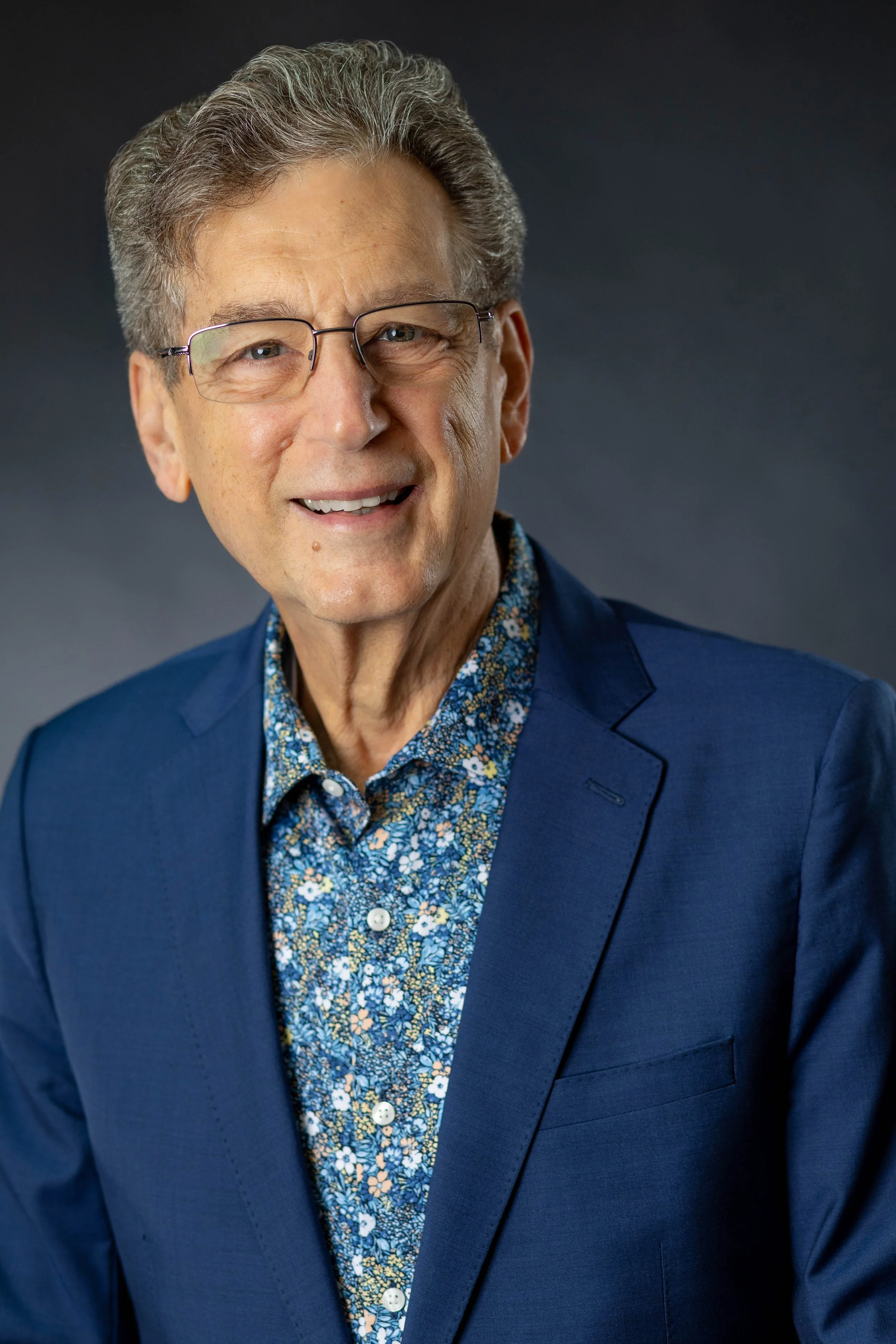 A smiling middle-aged man with gray hair, glasses, wearing a navy blazer over a patterned blue and white shirt, against a dark gray background.