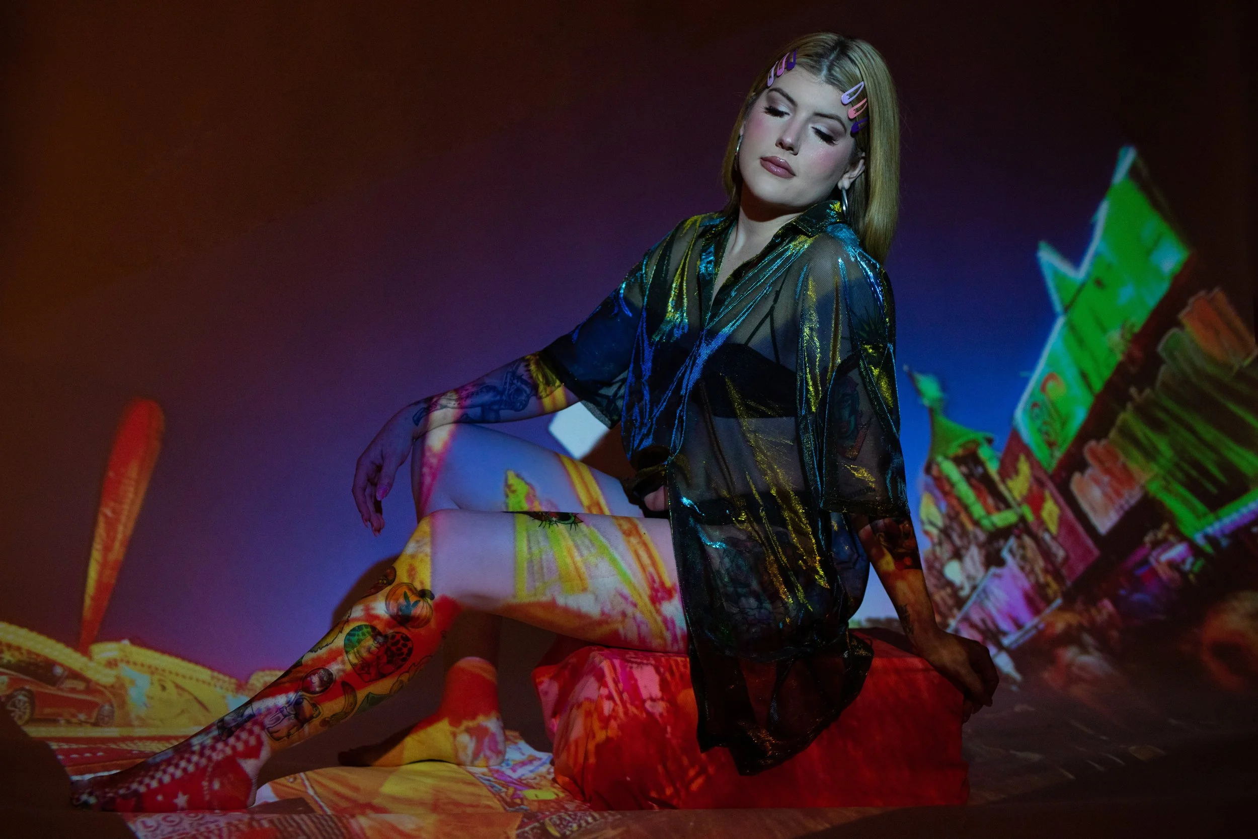 A woman with blonde hair and colorful hair clips sitting on a red cushion with her eyes closed, wearing a sheer black and gold top, with neon cityscape and colorful projections on her skin and clothes.