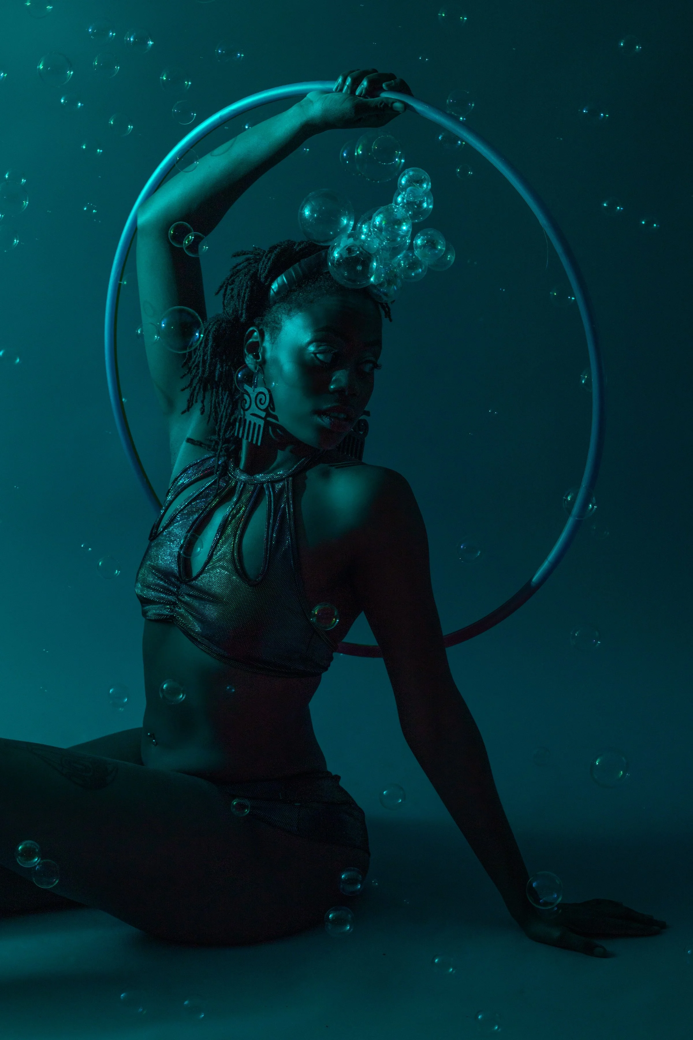 A woman underwater, wearing a mesh top and large earrings, poses with a floating hoop and bubbles around her.