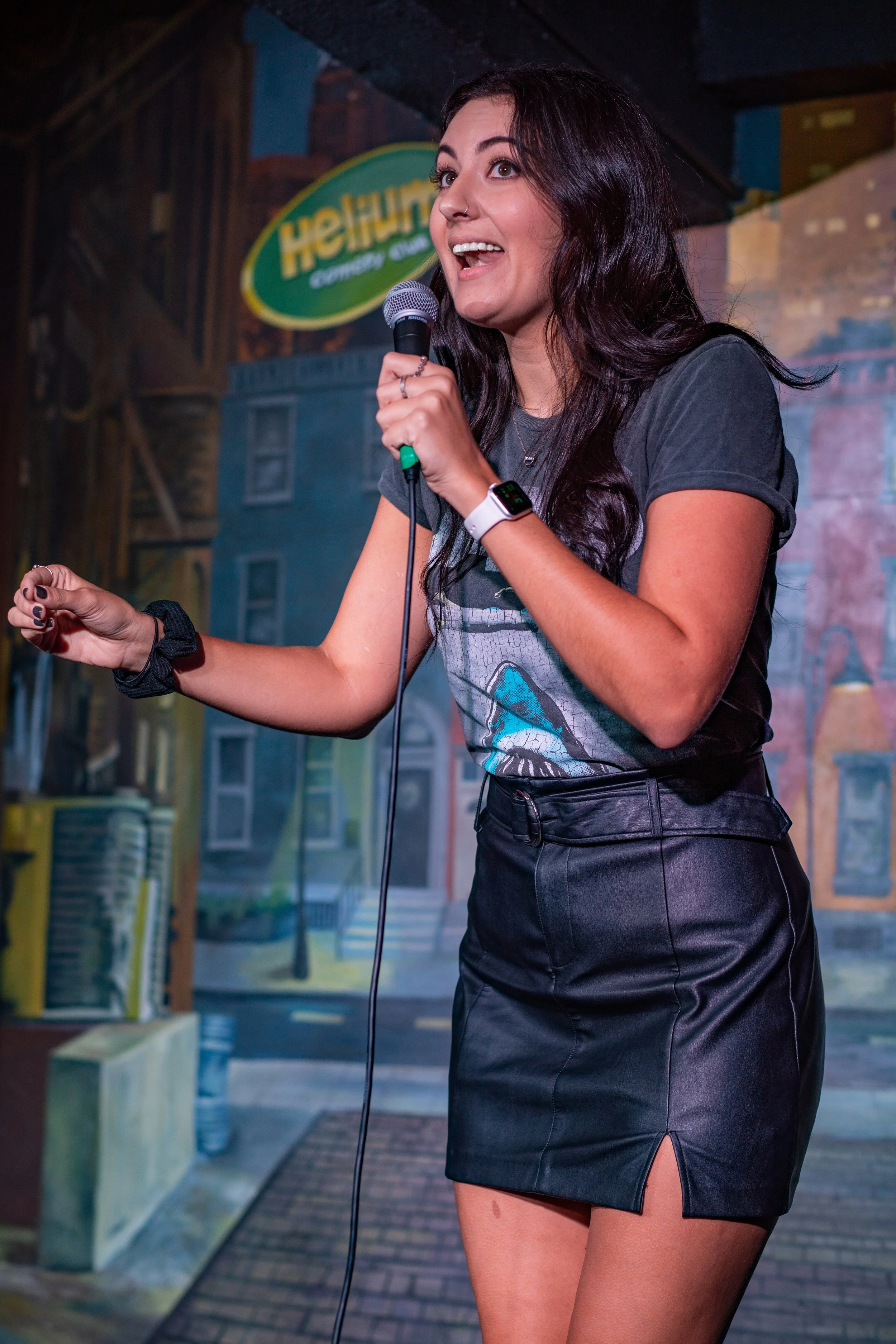 A woman performing stand-up comedy on stage with a microphone, background depicting a colorful cityscape mural.
