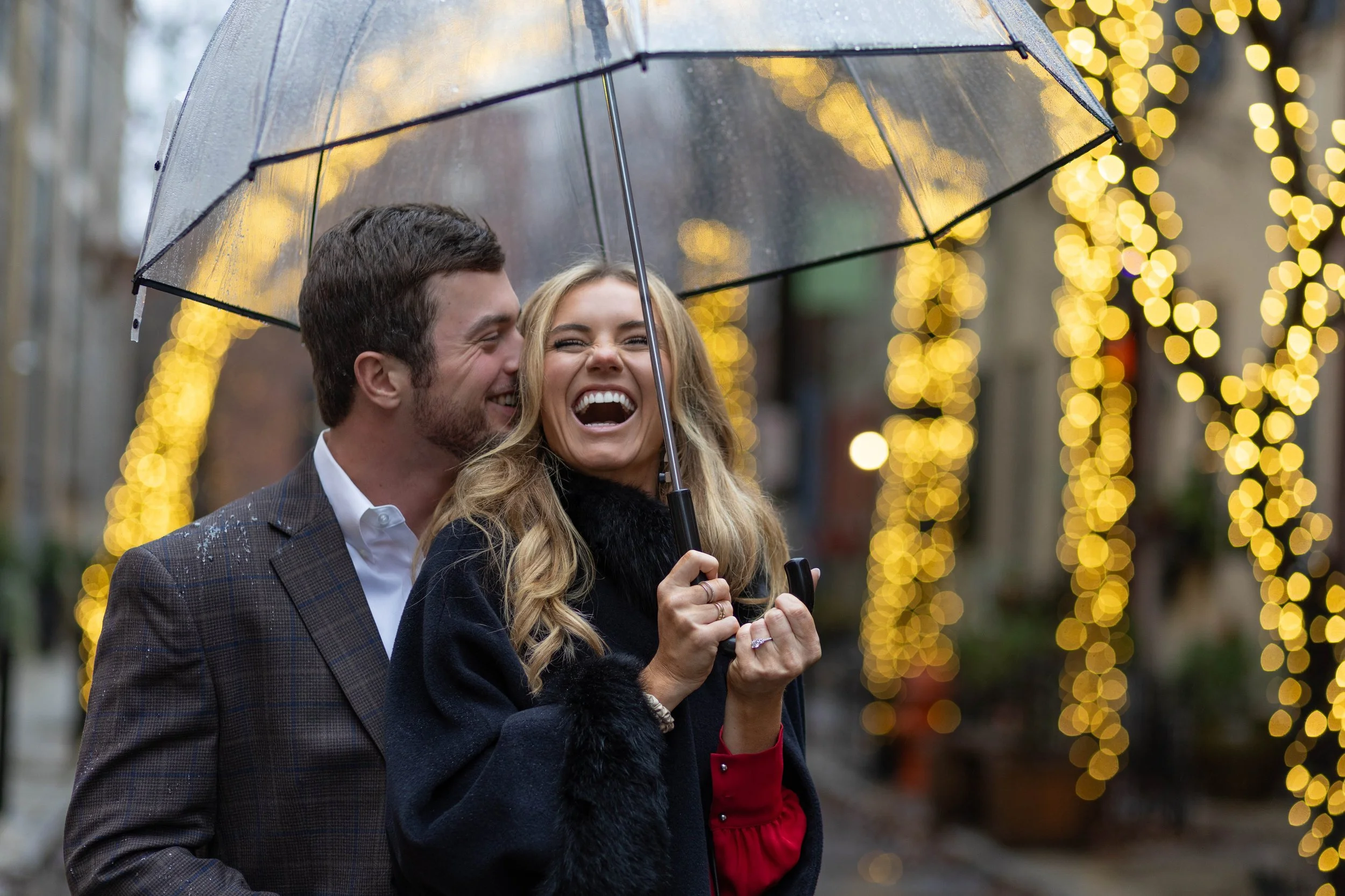 A joyful couple with an umbrella laughing, surrounded by festive Christmas lights on a rainy day.