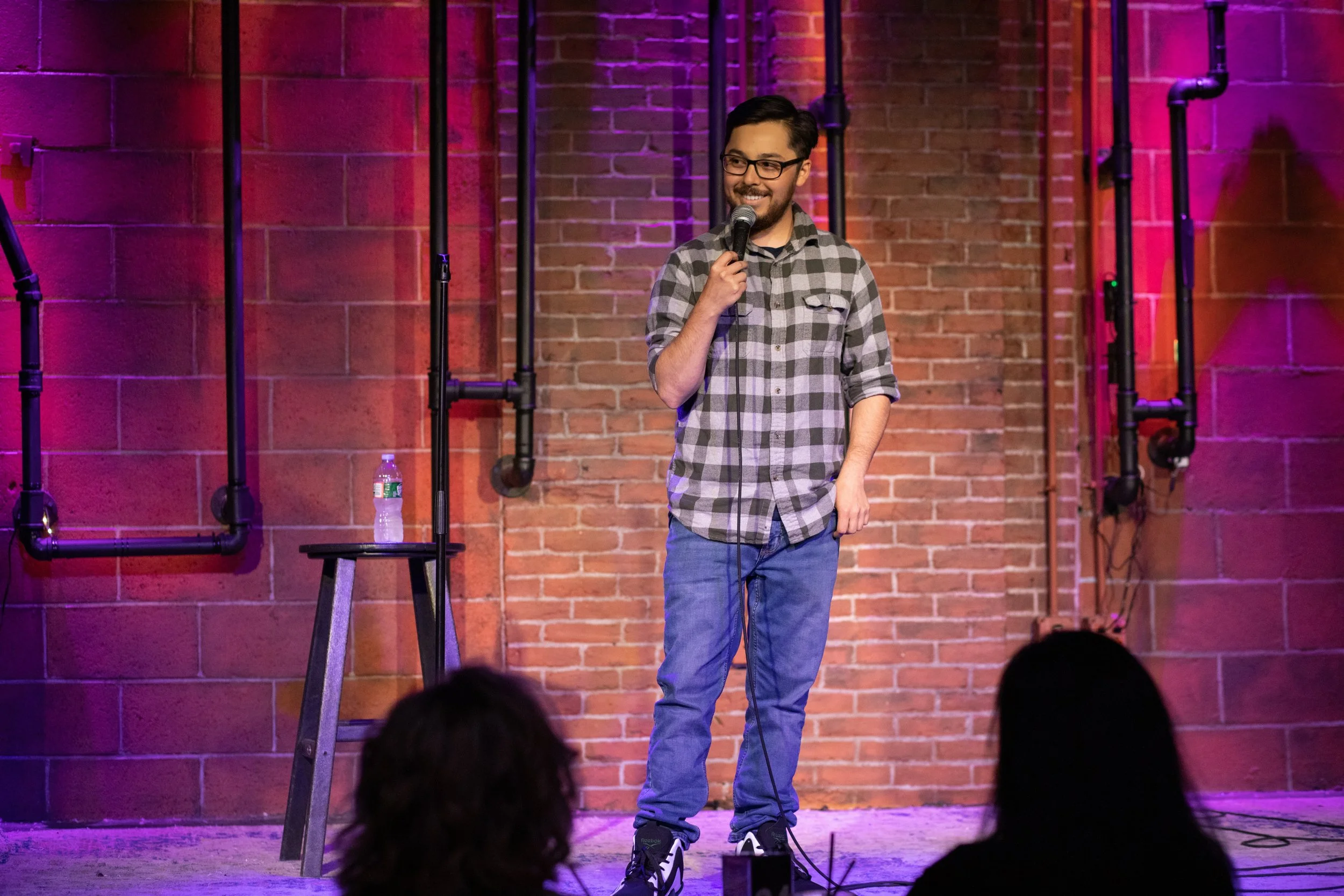 A male comedian standing on stage with a microphone, smiling, wearing a plaid shirt and jeans, in front of a brick wall with stage lighting.