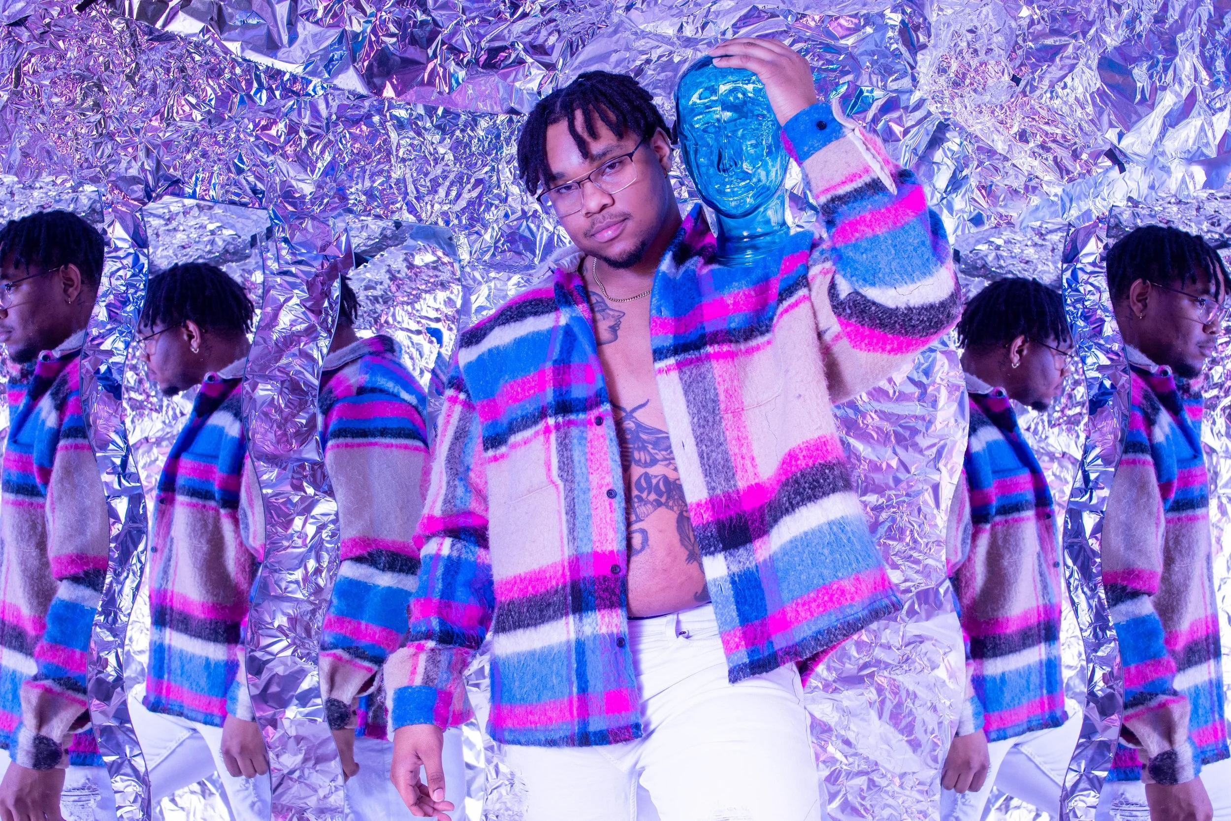 A young man with glasses and dreadlocks, shirtless with tattoos, wearing a colorful plaid jacket and white pants, holding a blue reflective mask, standing in front of multiple mirrors that reflect his image, against a metallic, crinkled silver backgr