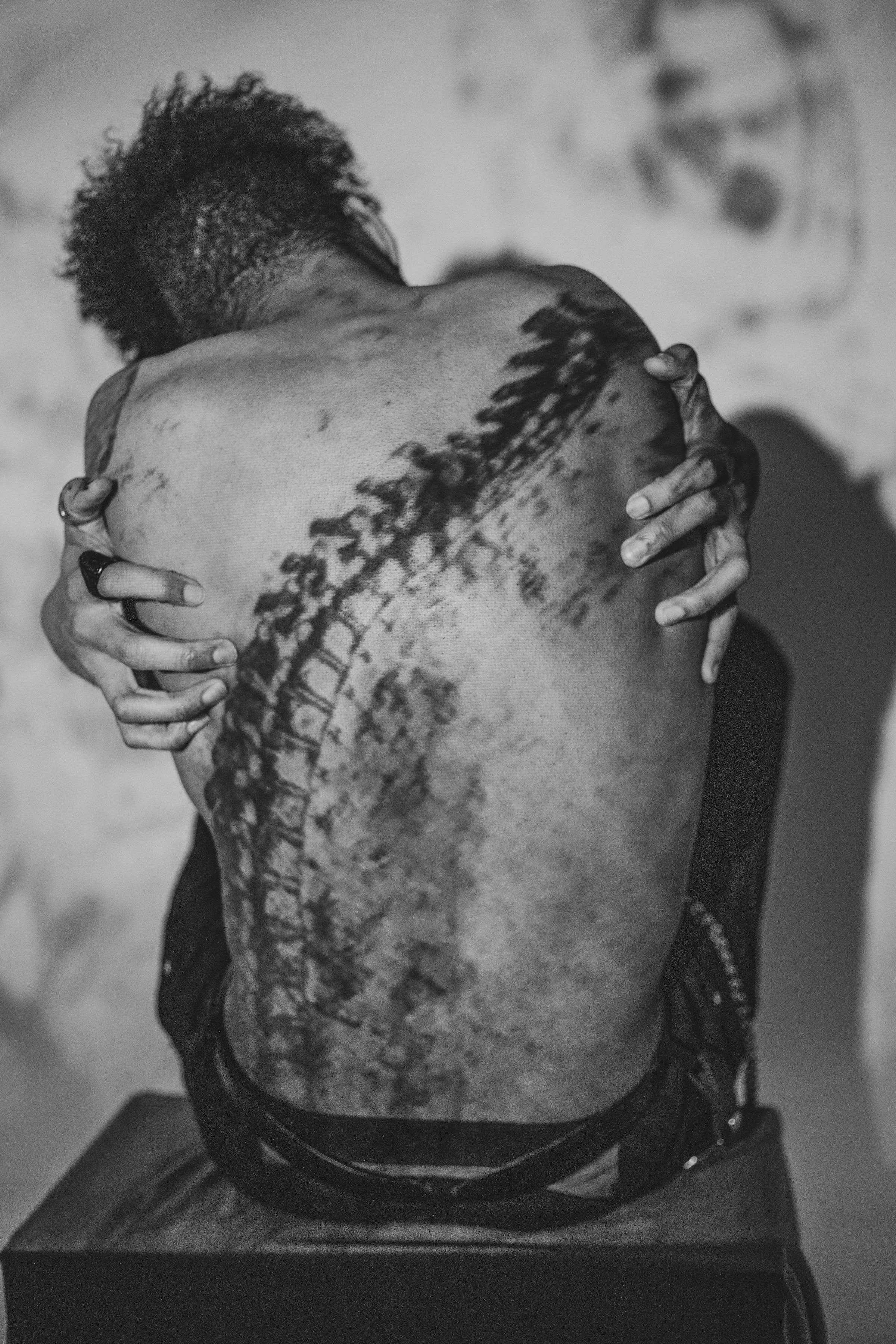 A person with short curly hair is facing away and hugging their own back, which has a large tattoo of a scenic mountain landscape with a bridge.