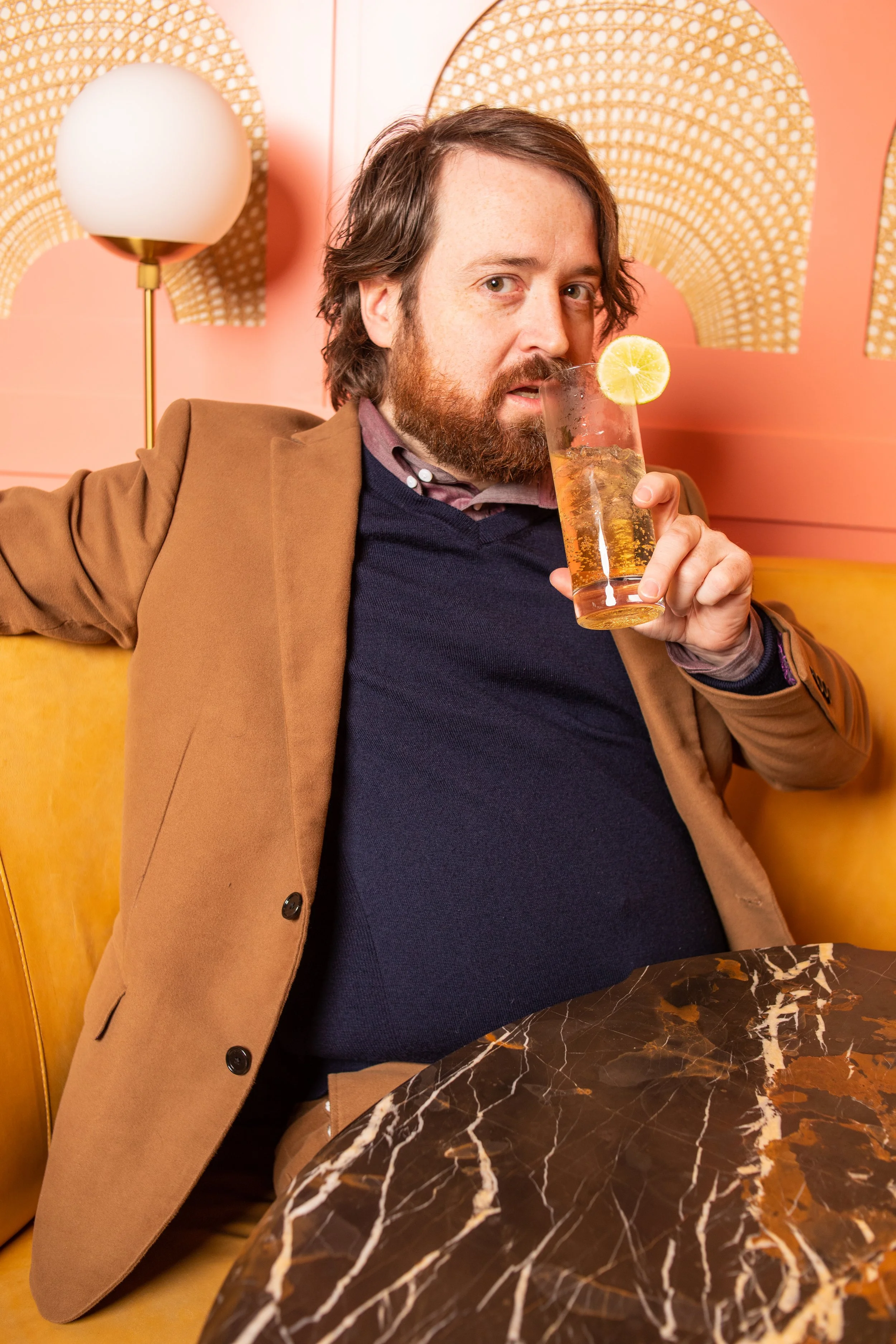 A man with a beard and long hair sitting in a retro-style booth, holding a glass of sparkling beverage with a lemon slice, wearing a brown blazer and navy sweater.