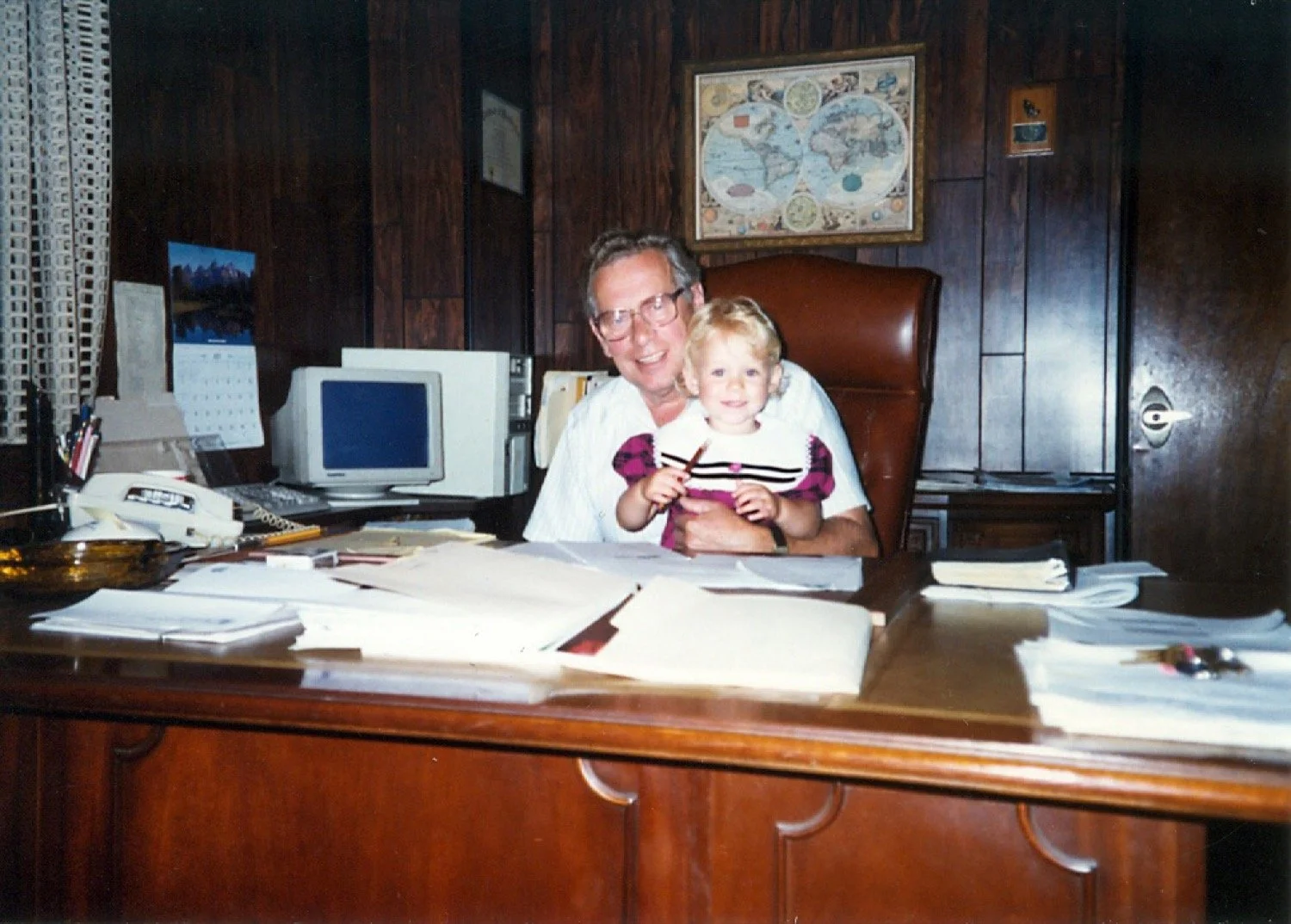 A man with glasses and a young girl sitting behind a cluttered office desk, smiling at the camera. The background features wood-paneled walls, framed maps, and office equipment.