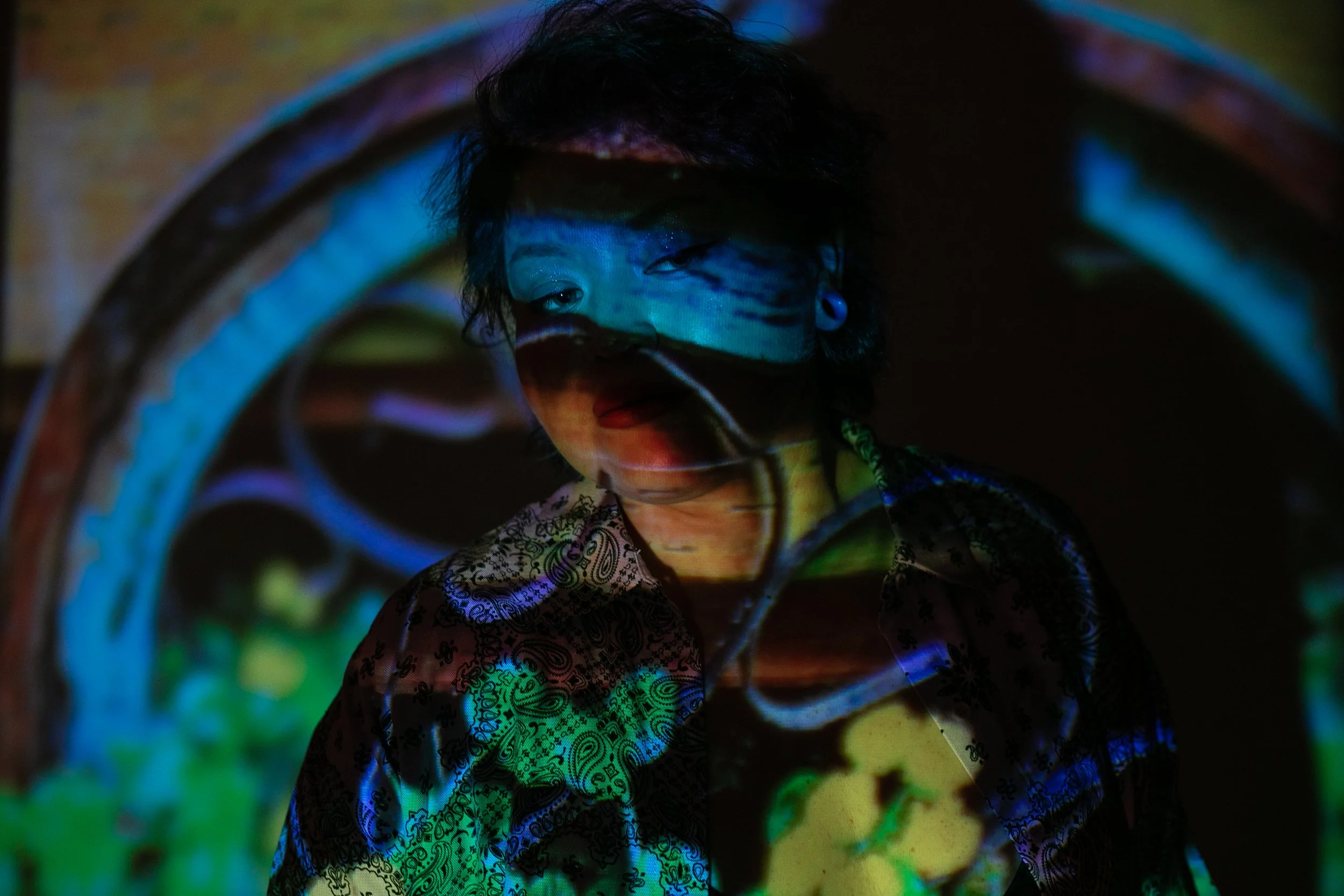 A young person with short, dark hair and piercings is seen through a circular metal arch, with colorful abstract patterns projected onto their face and clothing, creating a vibrant and artistic visual effect.