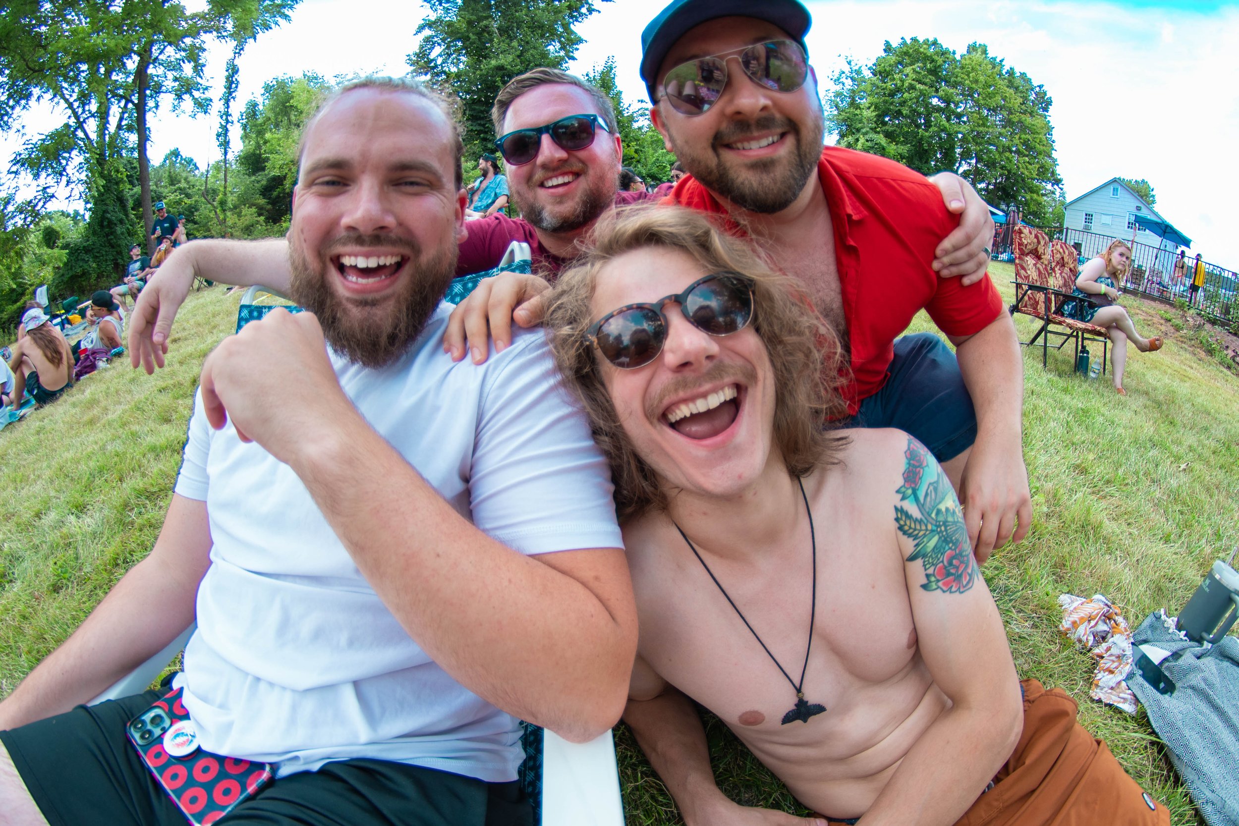 A group of five friends smiling and posing for a photo outdoors on a grassy area, with trees and other people in the background.