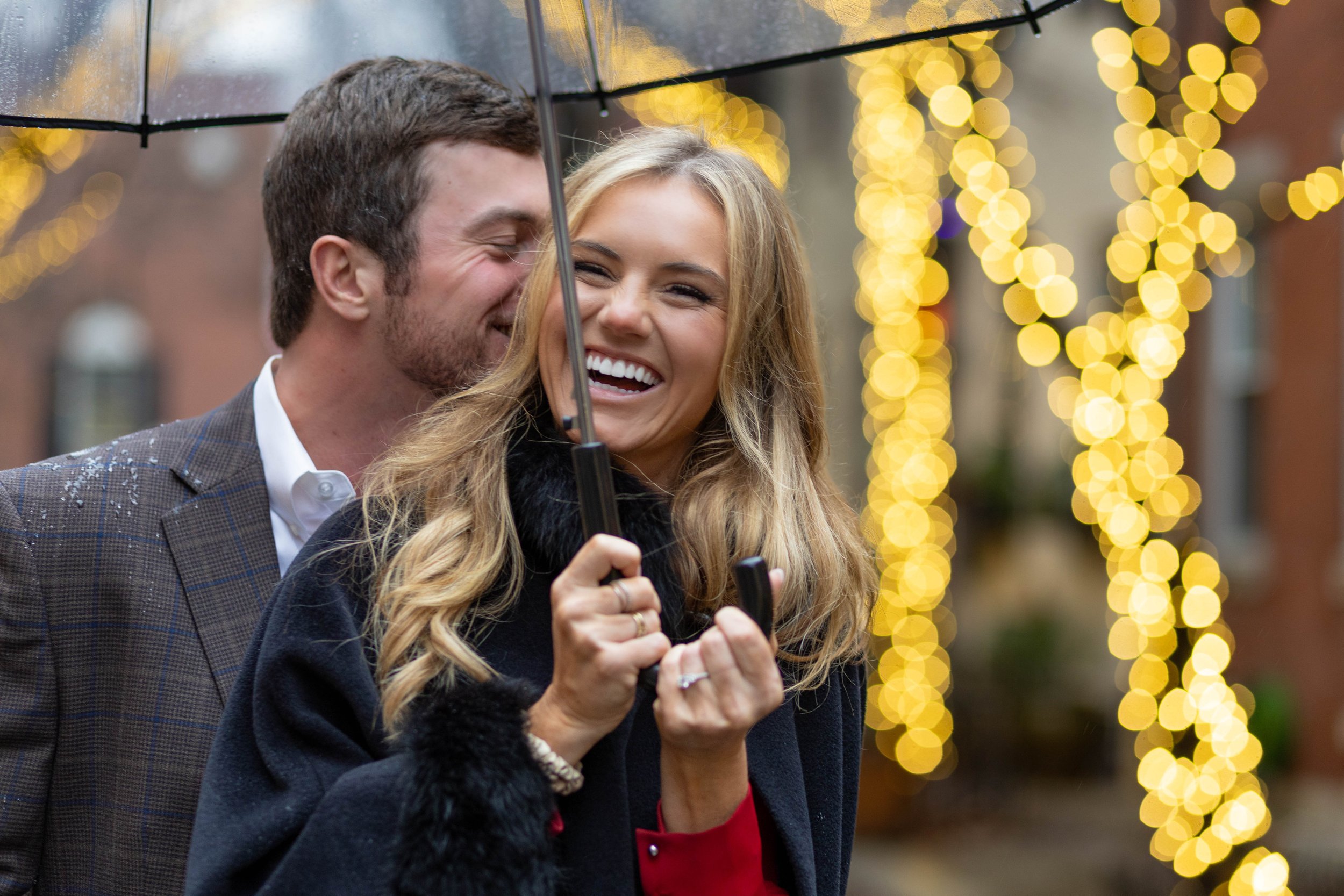 A smiling man and woman standing under an umbrella during a rainy evening, with blurred string lights in the background.