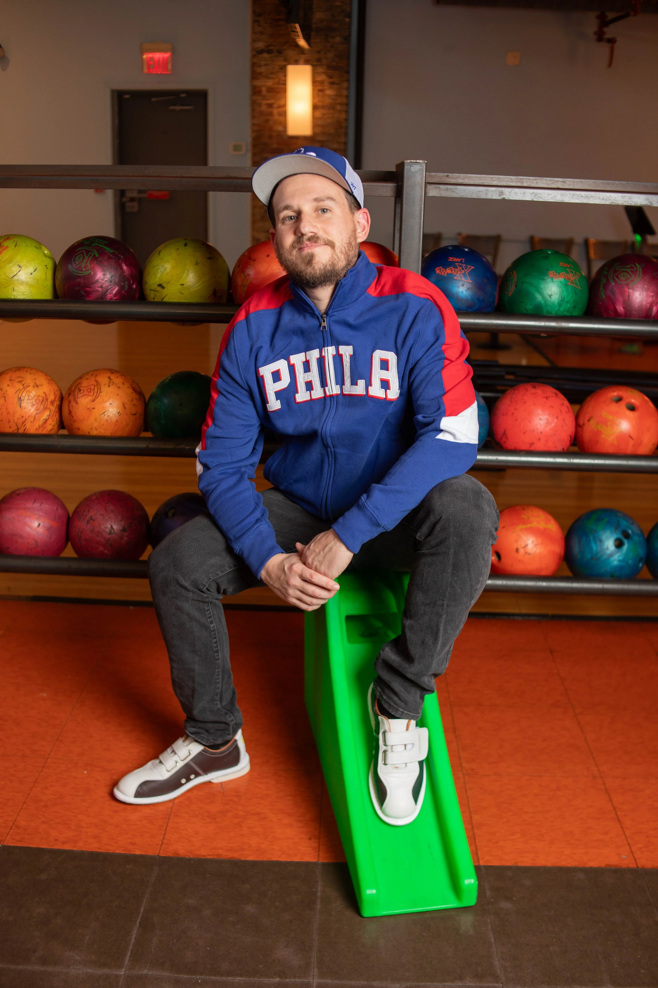 A man sitting on a green plastic slide in a bowling alley with a rack of colorful bowling balls behind him.