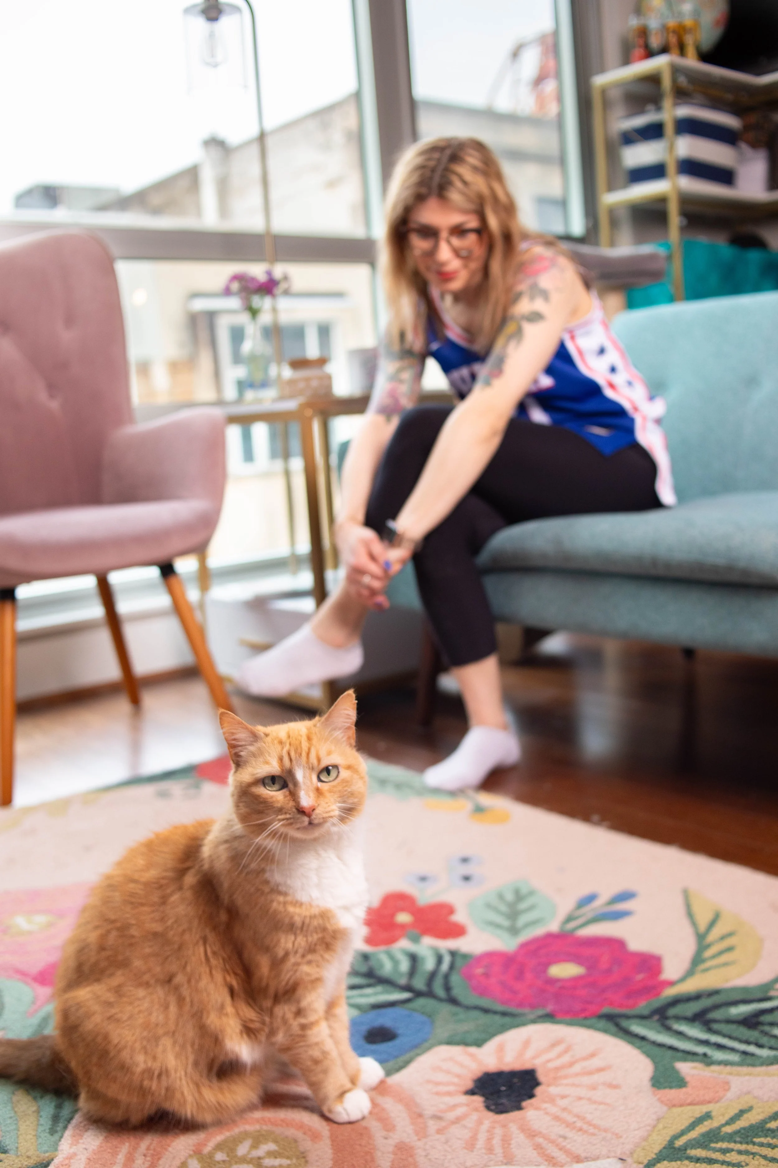 A woman with glasses and tattoos on her arms is sitting on a blue couch in a brightly lit room with large windows. She is leaning forward and adjusting her sock. In the foreground, an orange and white cat with green eyes is sitting on a colorful flor