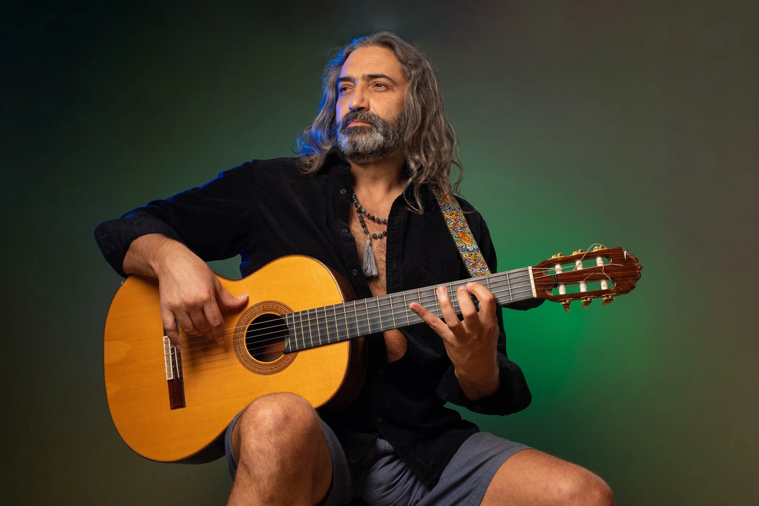 A man with long gray hair and a beard playing an acoustic guitar against a green background.