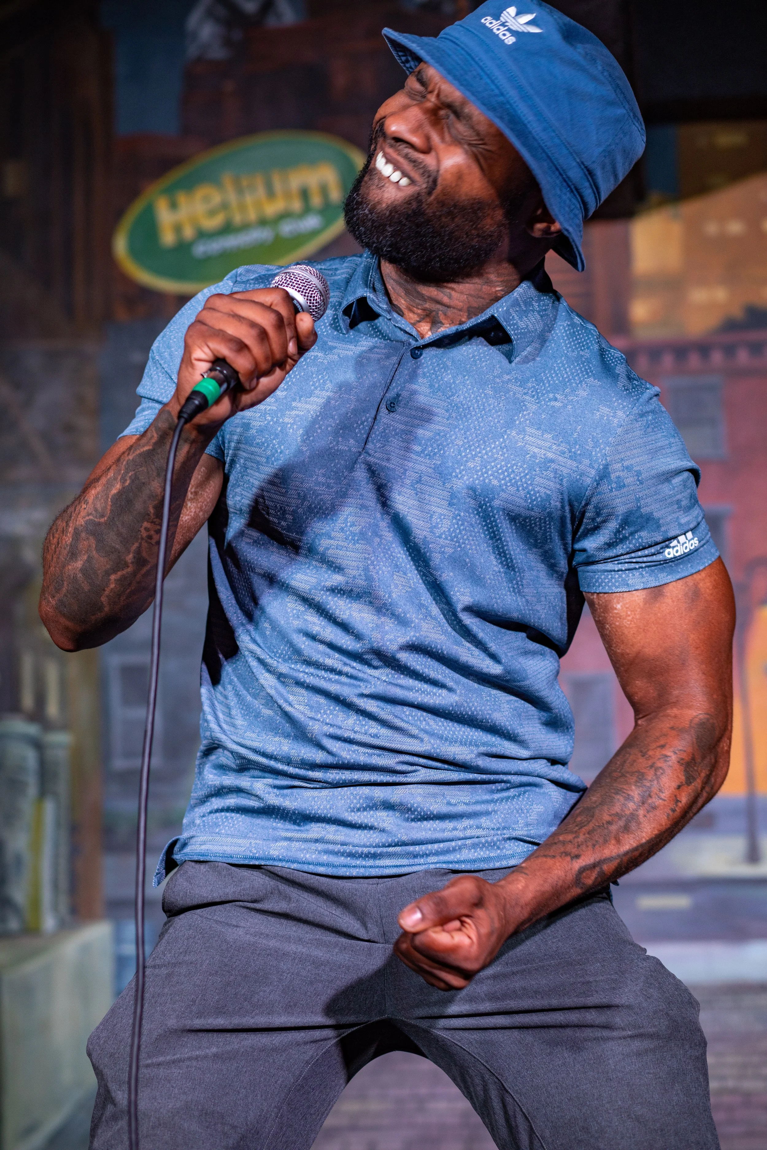 A man singing passionately into a microphone on stage, wearing a blue Adidas bucket hat and matching blue shirt, with tattoos on his arms. The background shows a sign and stage setup.