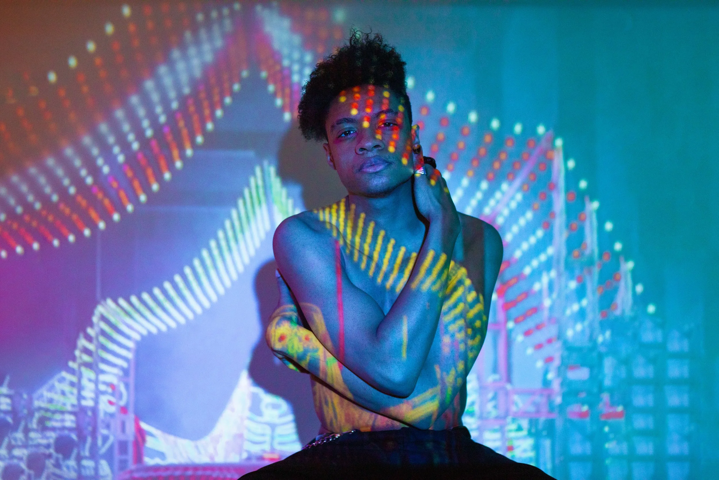 A topless person with short curly hair is posing with one hand on their cheek, surrounded by colorful, abstract light projections on the wall and their body.