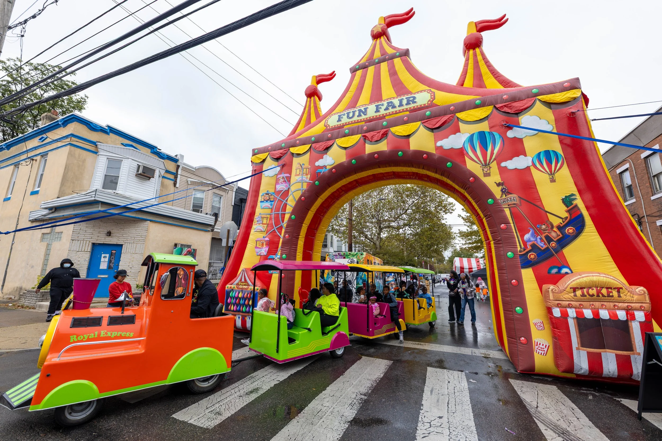 Colorful inflatable fun fair entrance arch with a small train ride in front and people around on a rainy day.