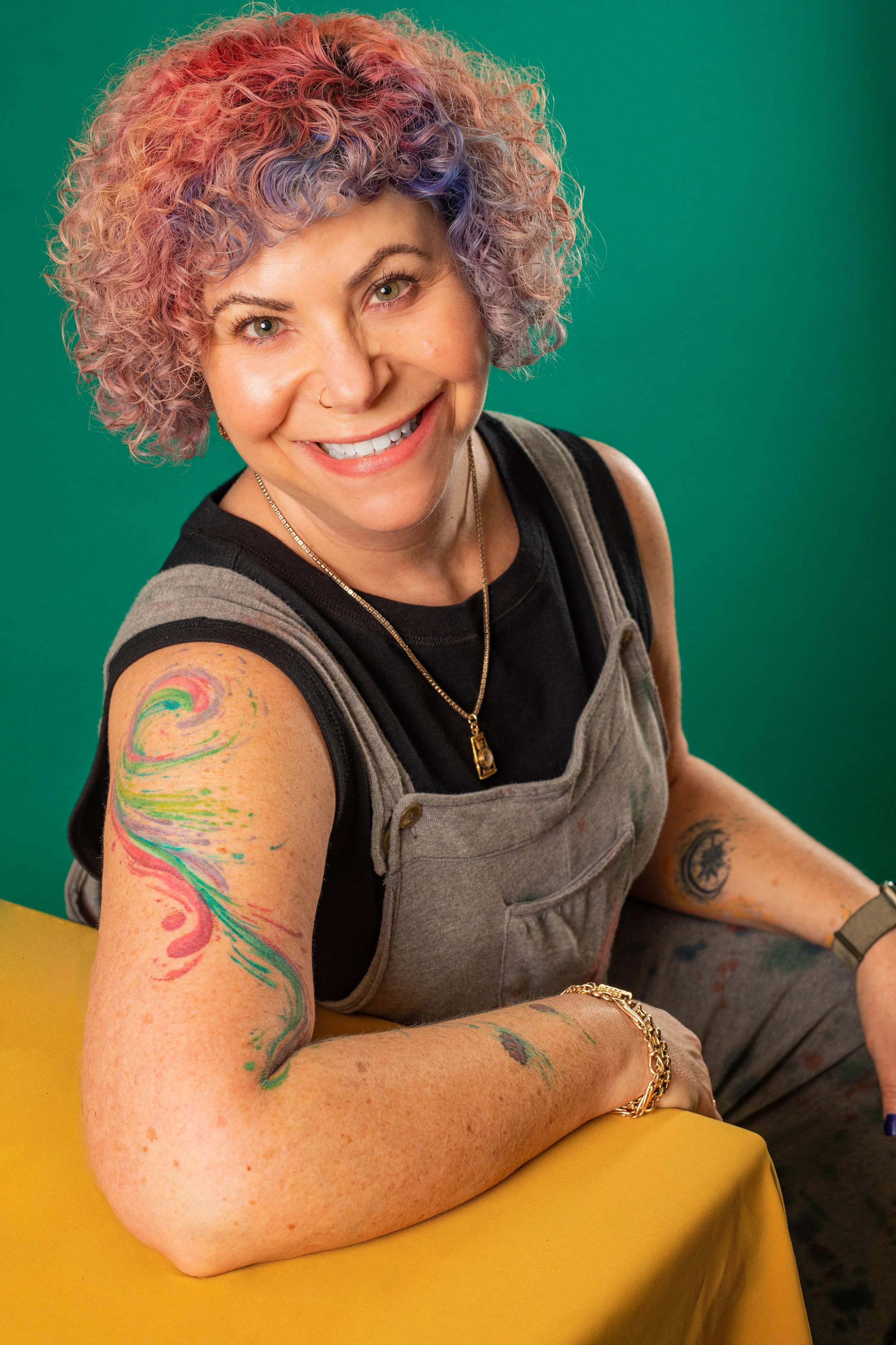 A woman with curly, colorful pastel hair, smiling at the camera, wearing a black shirt under gray overalls, with tattoos on her arms, a necklace, and a bracelet, sitting at a yellow table against a green background.