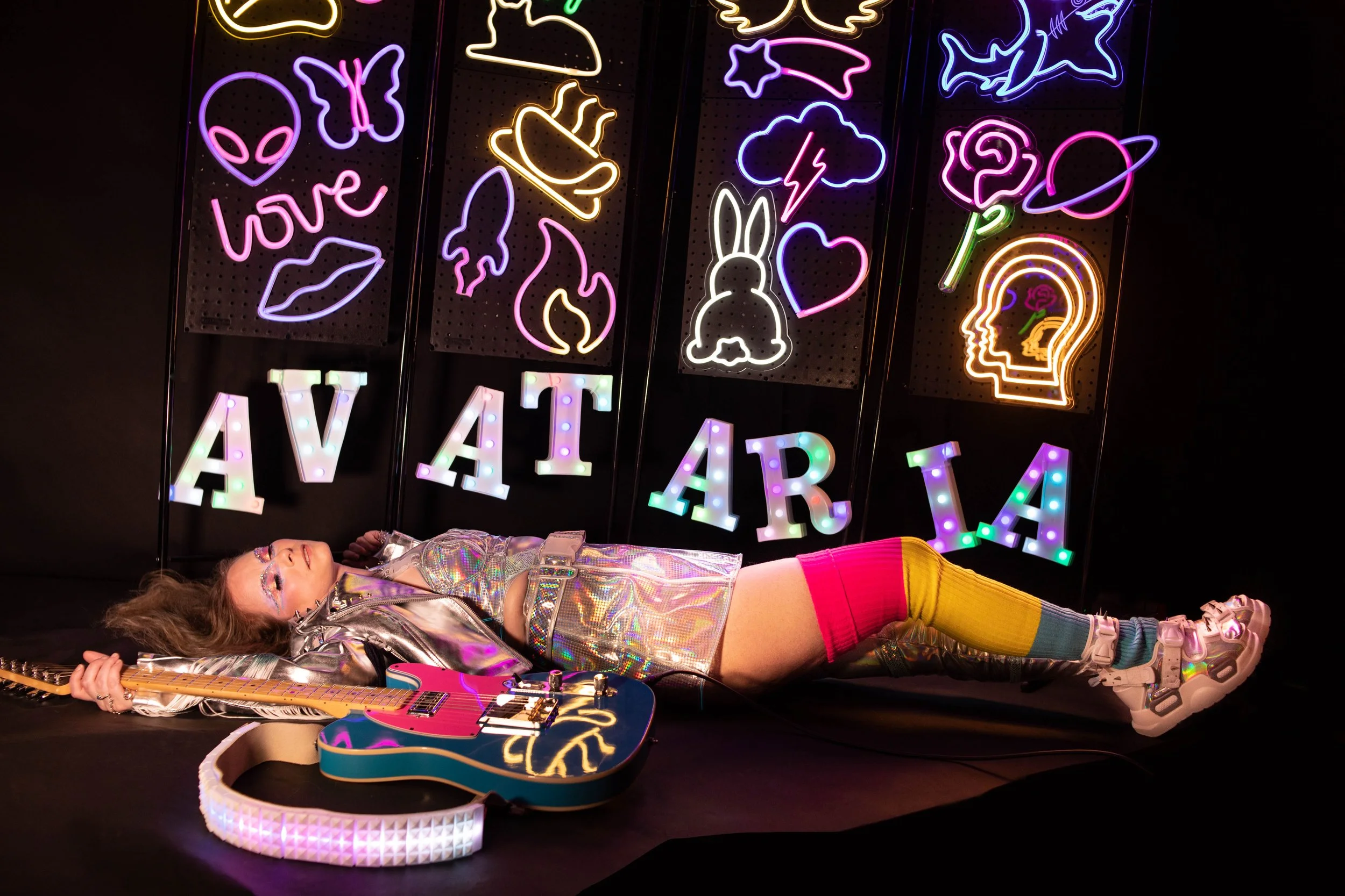 A person lying on the floor holding a guitar in front of a colorful neon sign wall. The person wears holographic clothing, multicolored leggings, and athletic shoes. The neon signs feature various symbols, including an alien head, butterfly, lips, bu