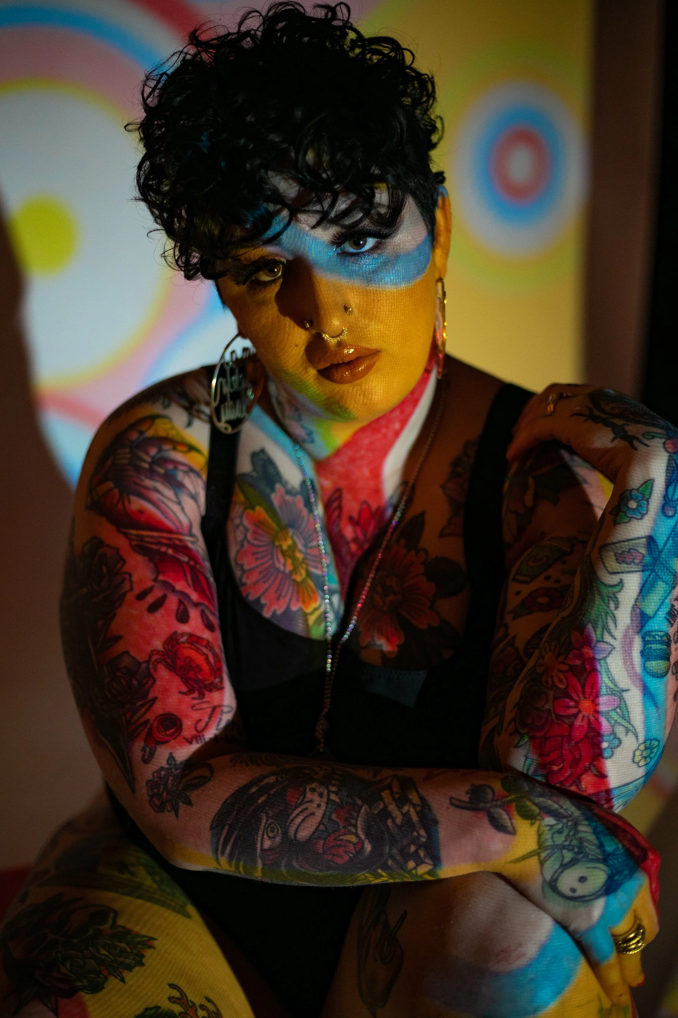 A person with tattoos on their arms and chest, wearing a black top, with Afro curly hair, face makeup, and jewelry, illuminated by colorful projections.