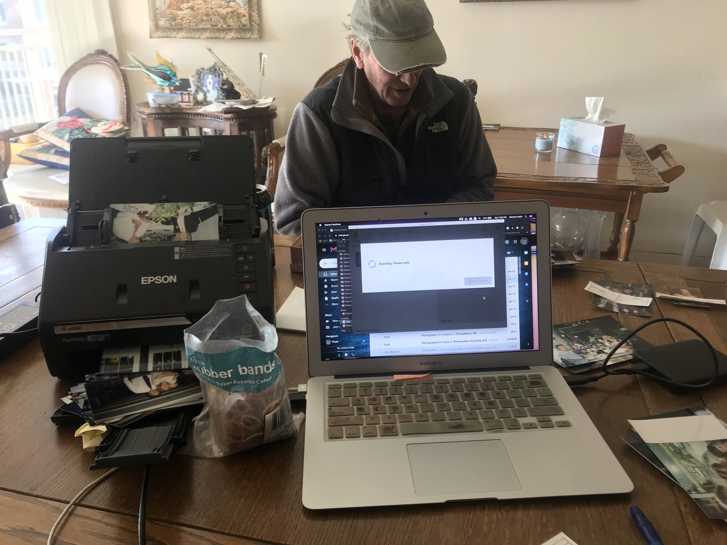 A man sitting at a cluttered wooden dining table with a laptop, an Epson scanner, and various photographs and papers. The man is wearing a cap and a North Face jacket and is looking down. The laptop screen shows a scanning interface, and there are other items like a bag of rubber bands and a pen on the table.