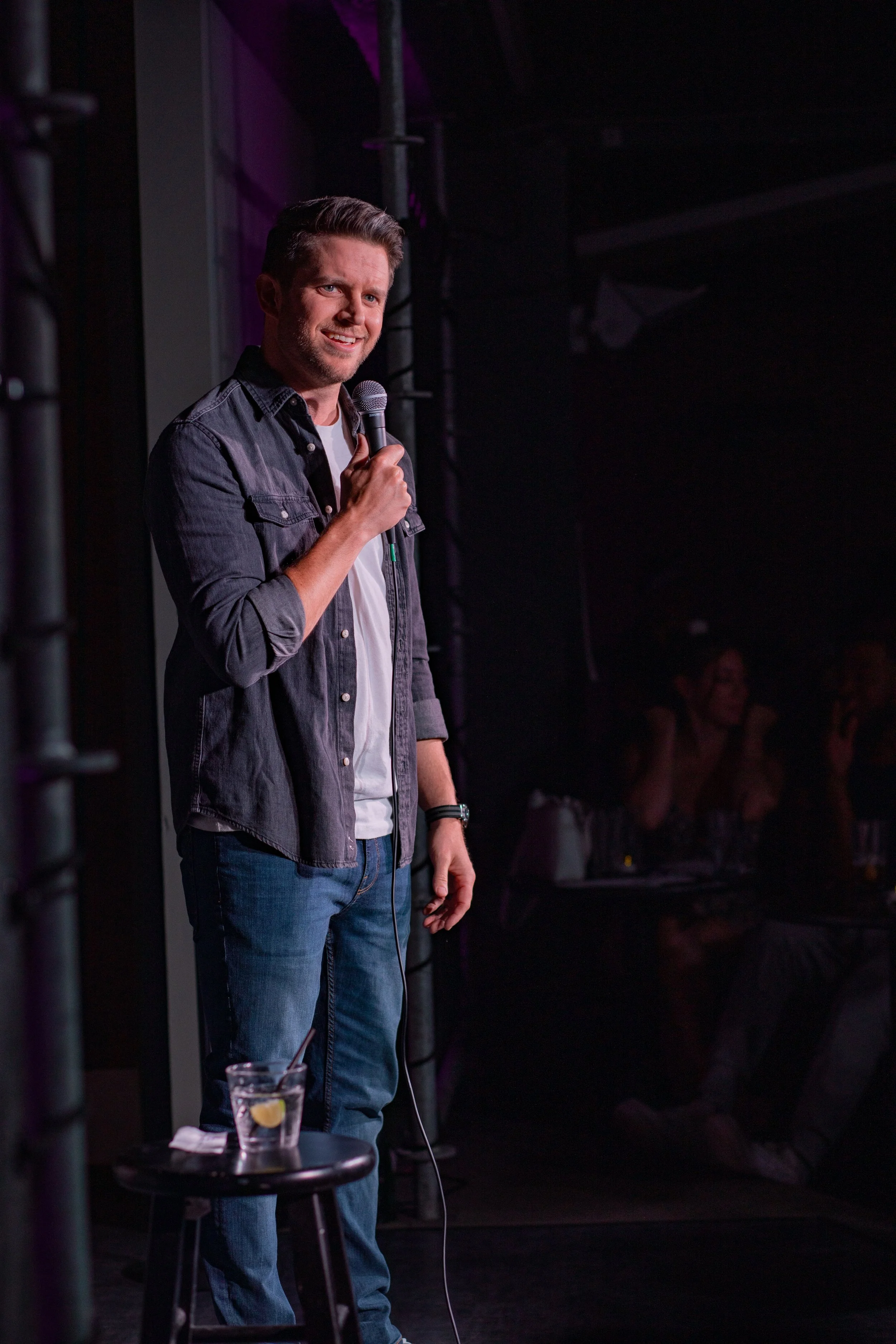 Stand-up comedian on stage with microphone, performing in front of audience, audience members smiling and watching, drink with lemon on stool.