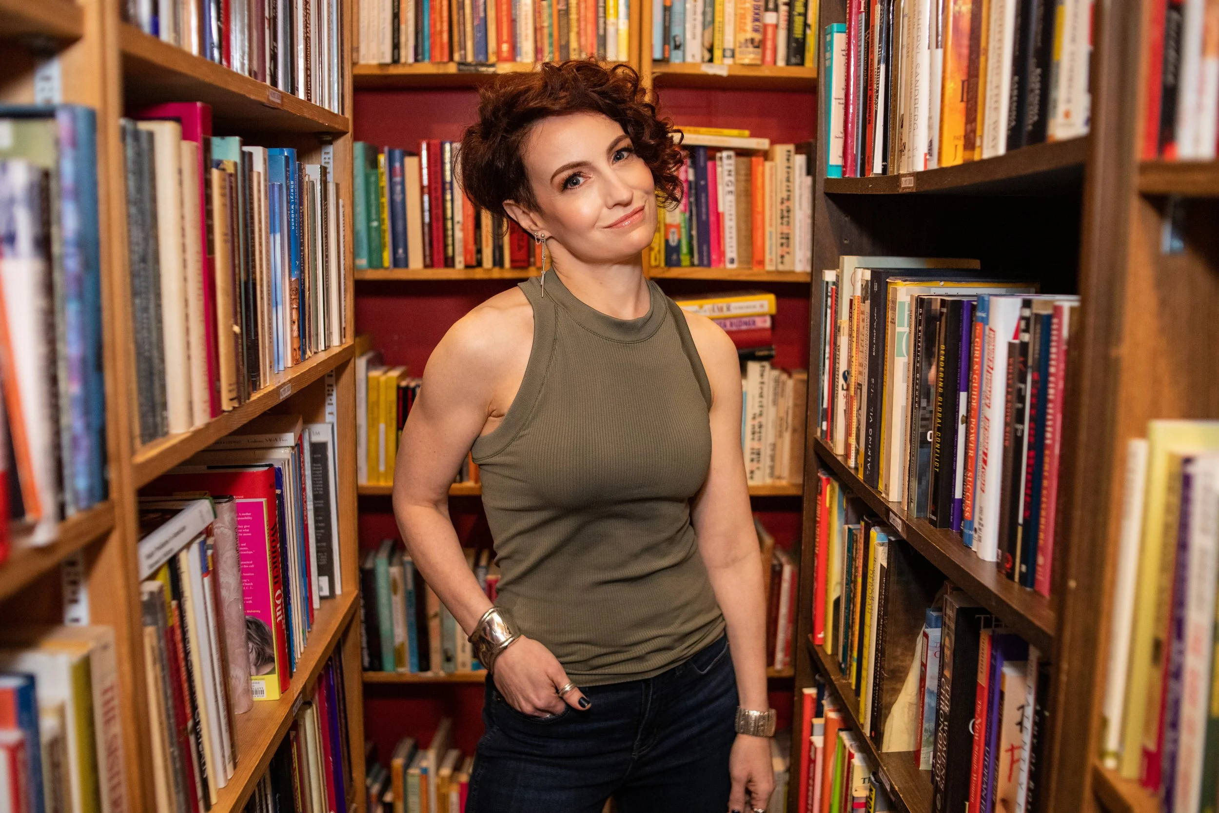 A woman with short curly brown hair, wearing a sleeveless olive green top, standing between bookshelves filled with colorful books in a library or bookstore.