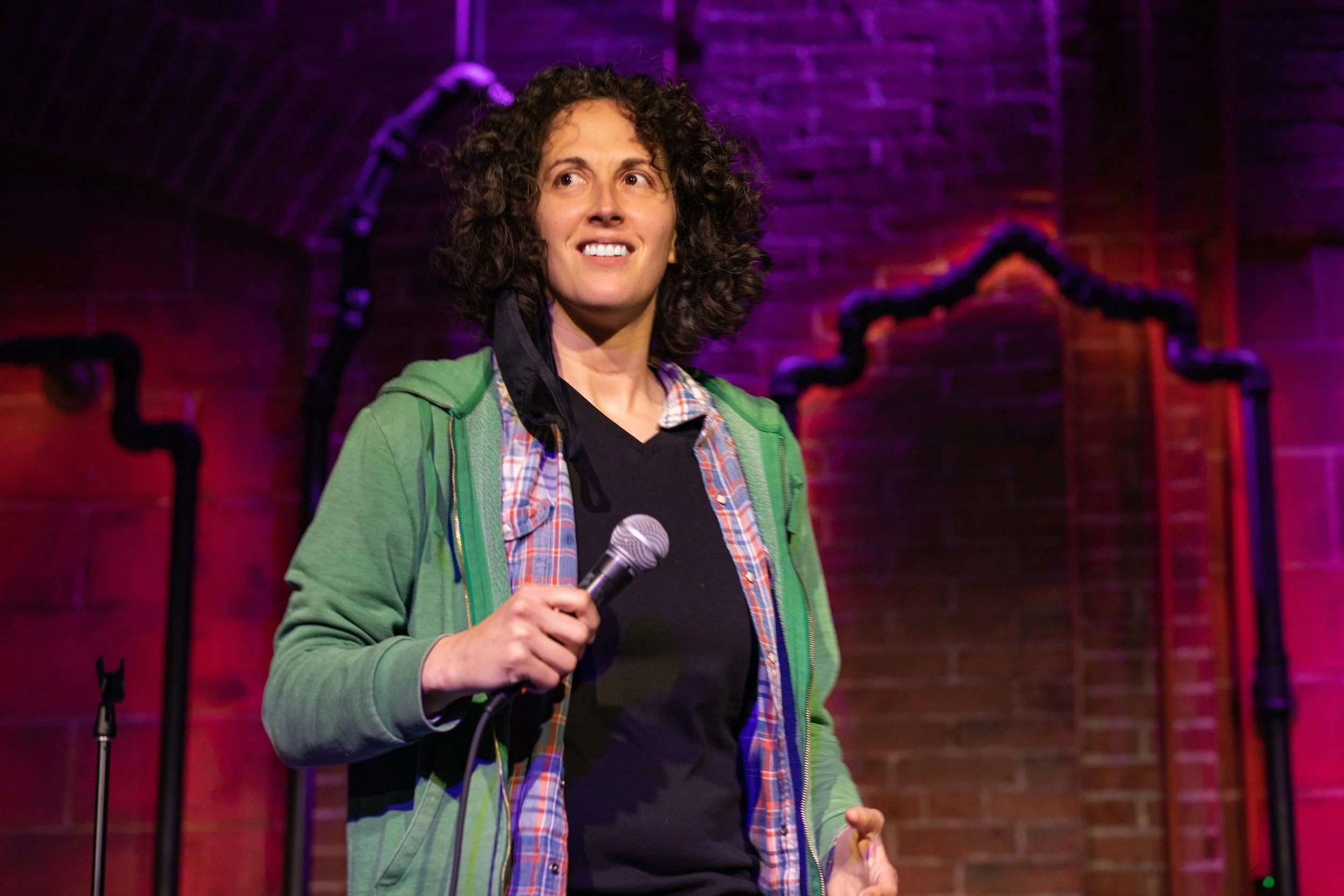 A woman with curly brown hair holds a microphone on stage, standing in front of a brick wall with purple and pink stage lighting.