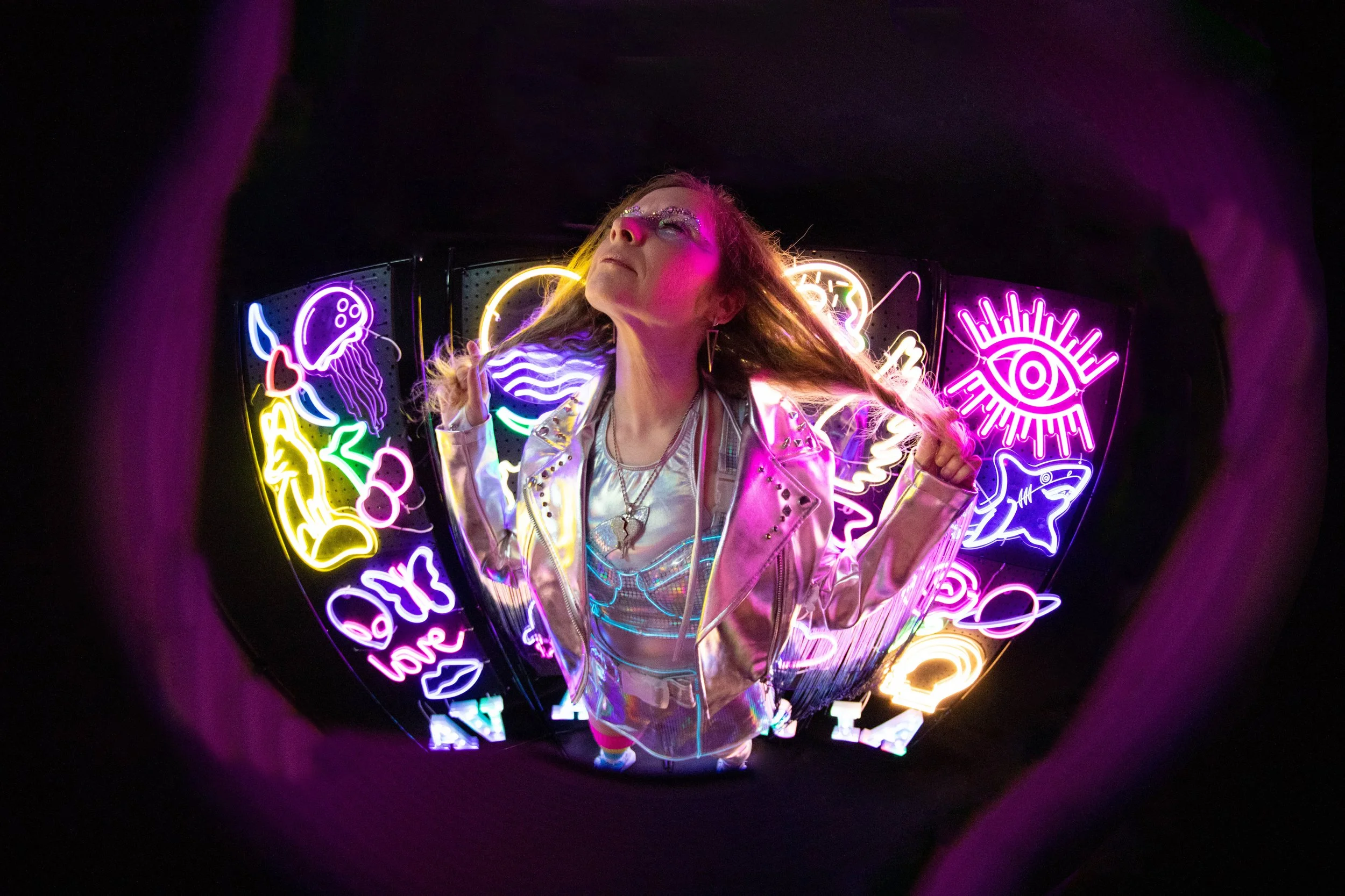 A woman with long hair and glittery makeup, wearing a shiny metallic jacket and necklaces, stands in front of a black background filled with colorful neon signs, including a jellyfish, an eye, a fish, and hearts, with her eyes closed and her head til