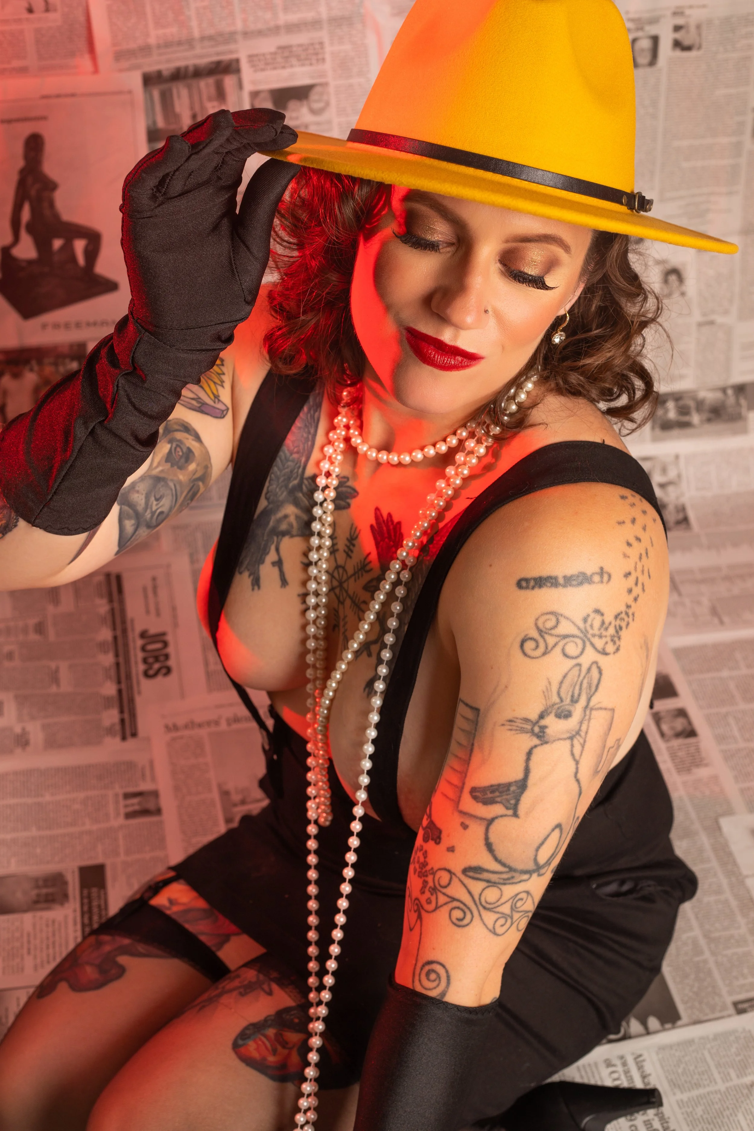 A woman with tattoos and red lipstick, wearing a yellow hat, black dress, and long black gloves, poses seductively in front of a backdrop of newspapers. She is accessorized with pearl necklaces and is holding the brim of her hat.