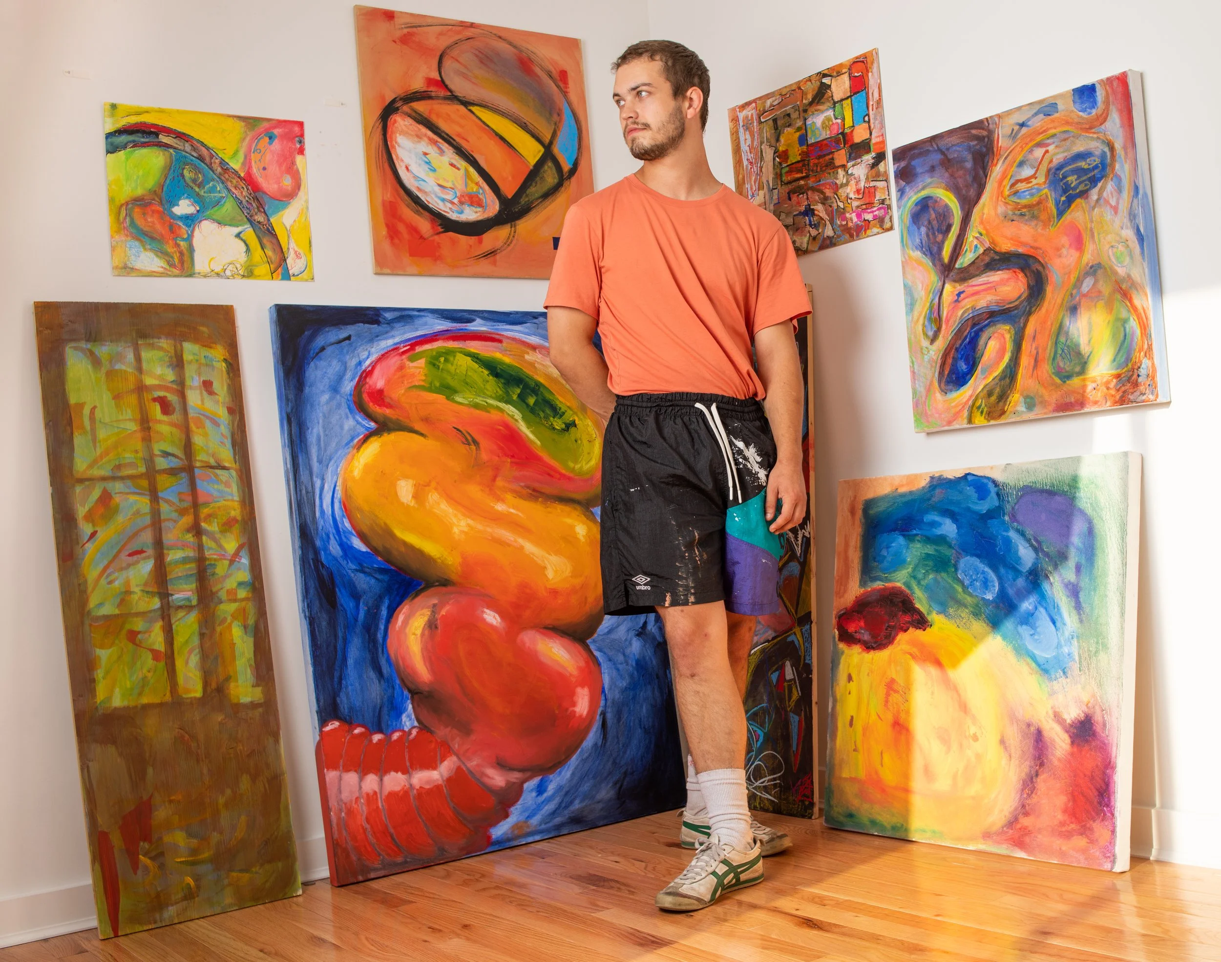 A young man stands in an art gallery surrounded by colorful abstract paintings. He is wearing an orange t-shirt, black shorts with paint splatters, white socks, and sneakers, and looks to the side.