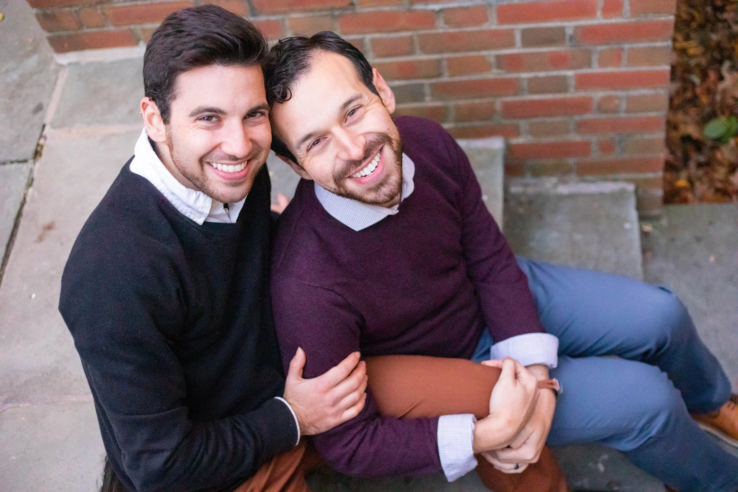 Two smiling men sitting on a step against a brick wall, embracing each other