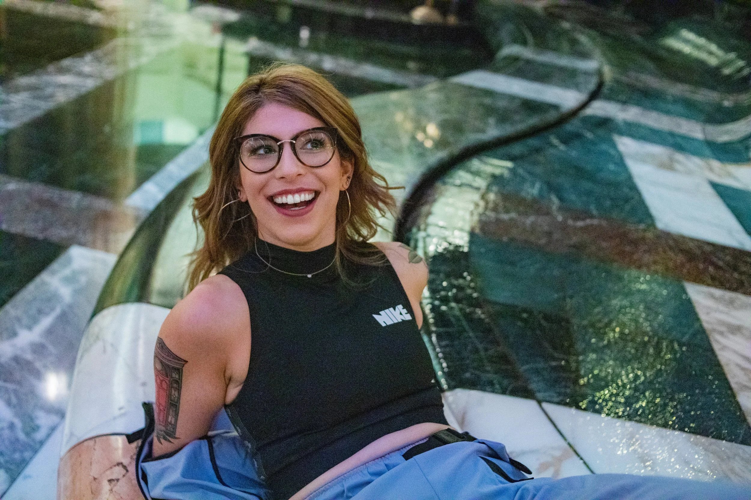 A young woman with glasses, tattoos, and a black Nike tank top is sitting on a white float in a pool. She is smiling and looking up, with water and pool tiles visible in the background.