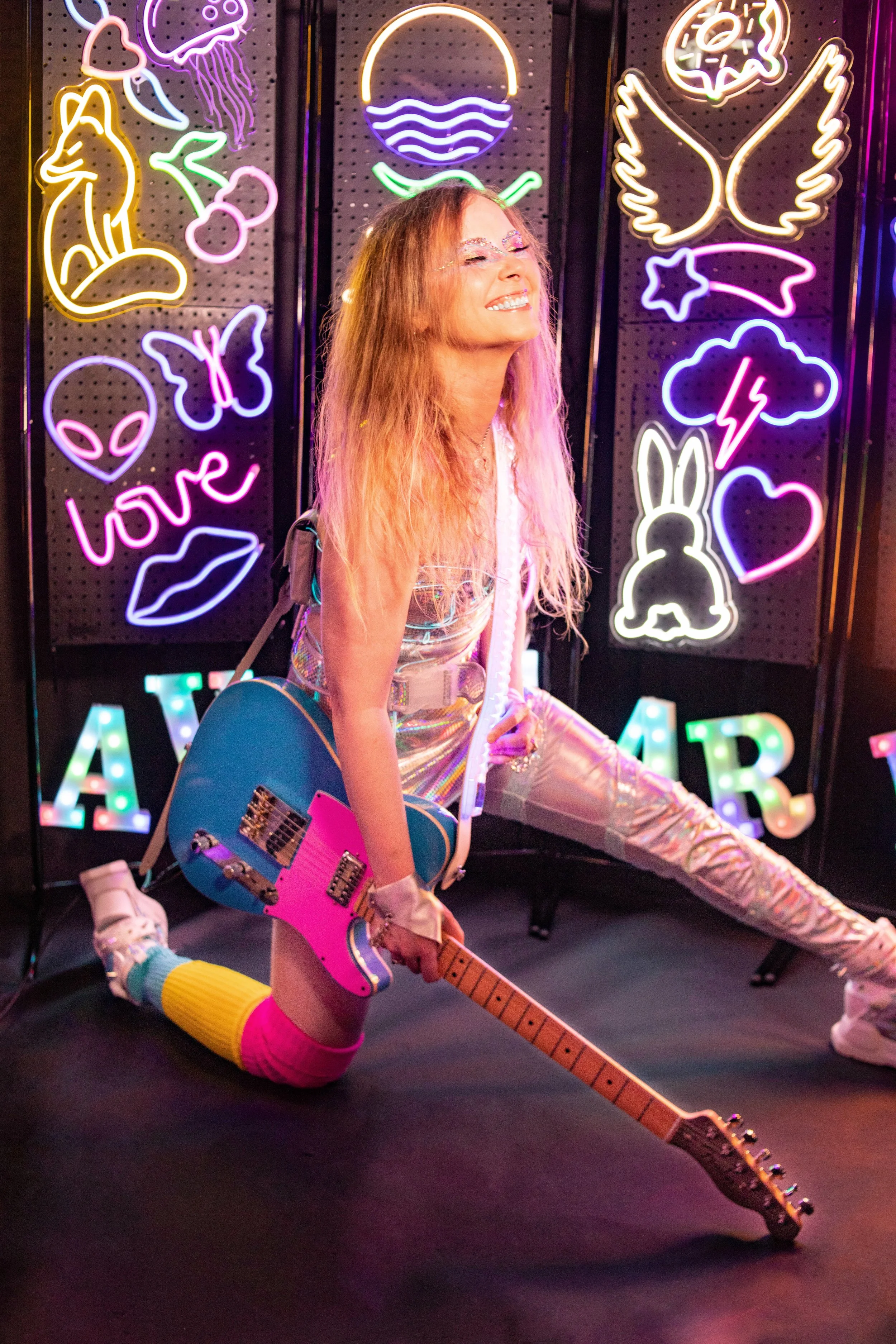 A woman with wavy, colorful hair kneels on the floor, holding a blue electric guitar. She is smiling with her eyes closed, wearing holographic pants, a clear top, and colorful socks. Behind her, there are vibrant neon signs of various shapes, includi