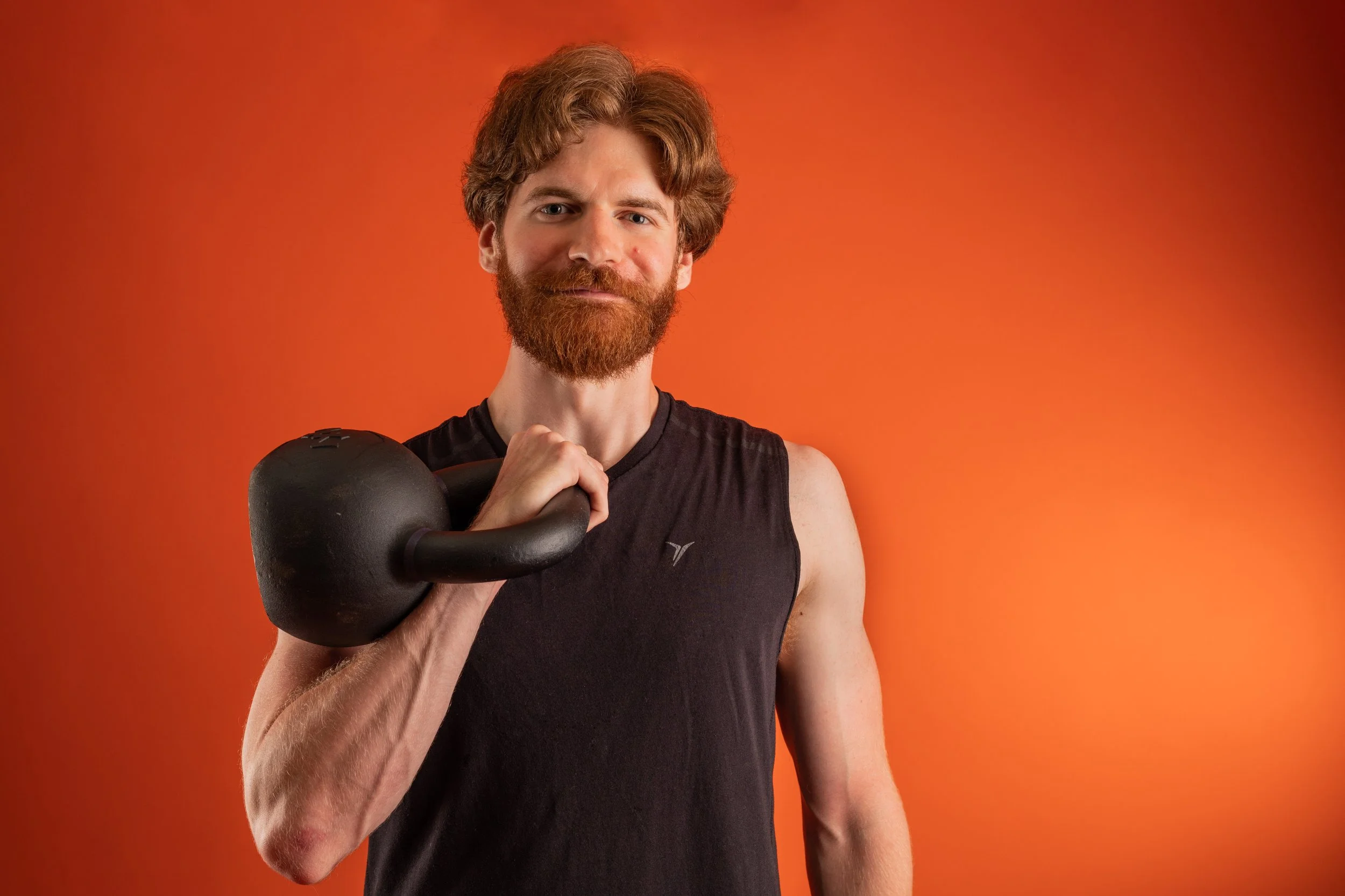 A man with a red beard and wavy hair holding a black kettlebell over his shoulder, wearing a black sleeveless athletic shirt, standing against an orange background.