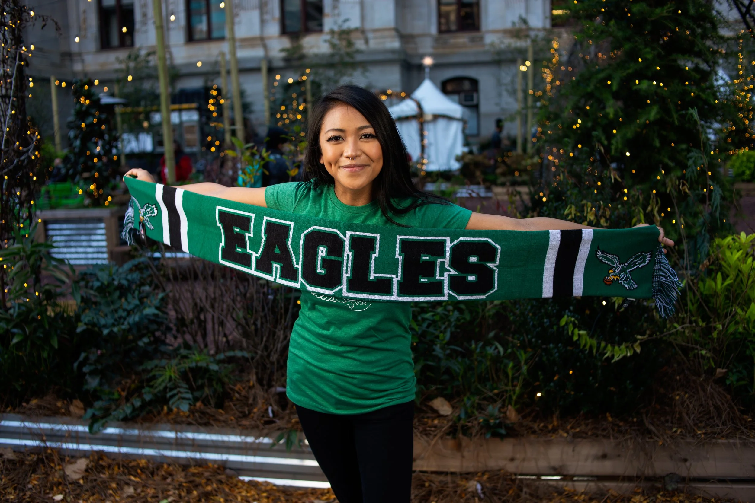 A woman in a green shirt holds a green Eagles scarf with both arms extended, smiling at the camera, outdoors in a garden area with decorative lights and buildings in the background.
