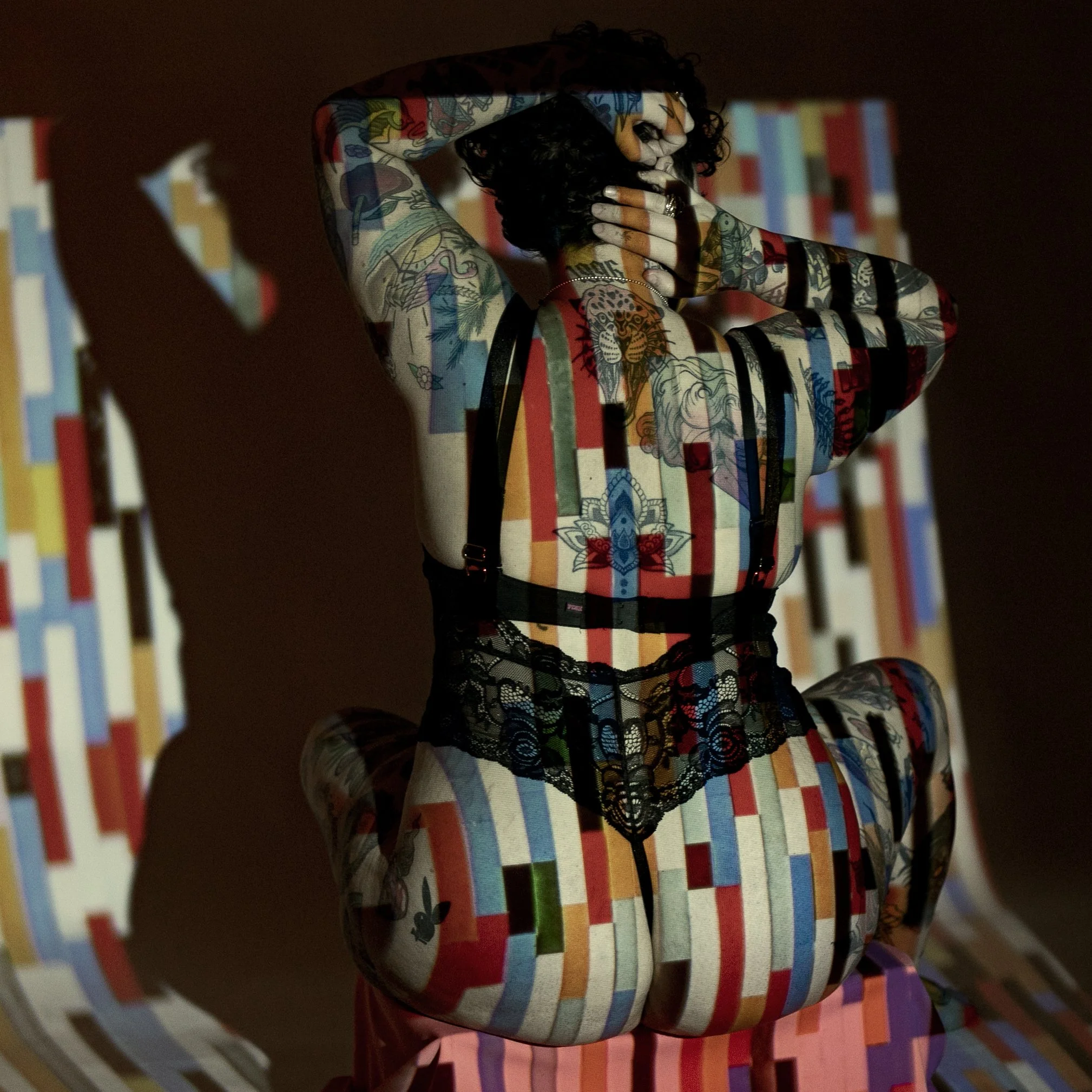 A woman with tattoos, black hair, and a nose ring posed with her hands on her head, with a colorful, pixelated pattern projected across her body and the background.