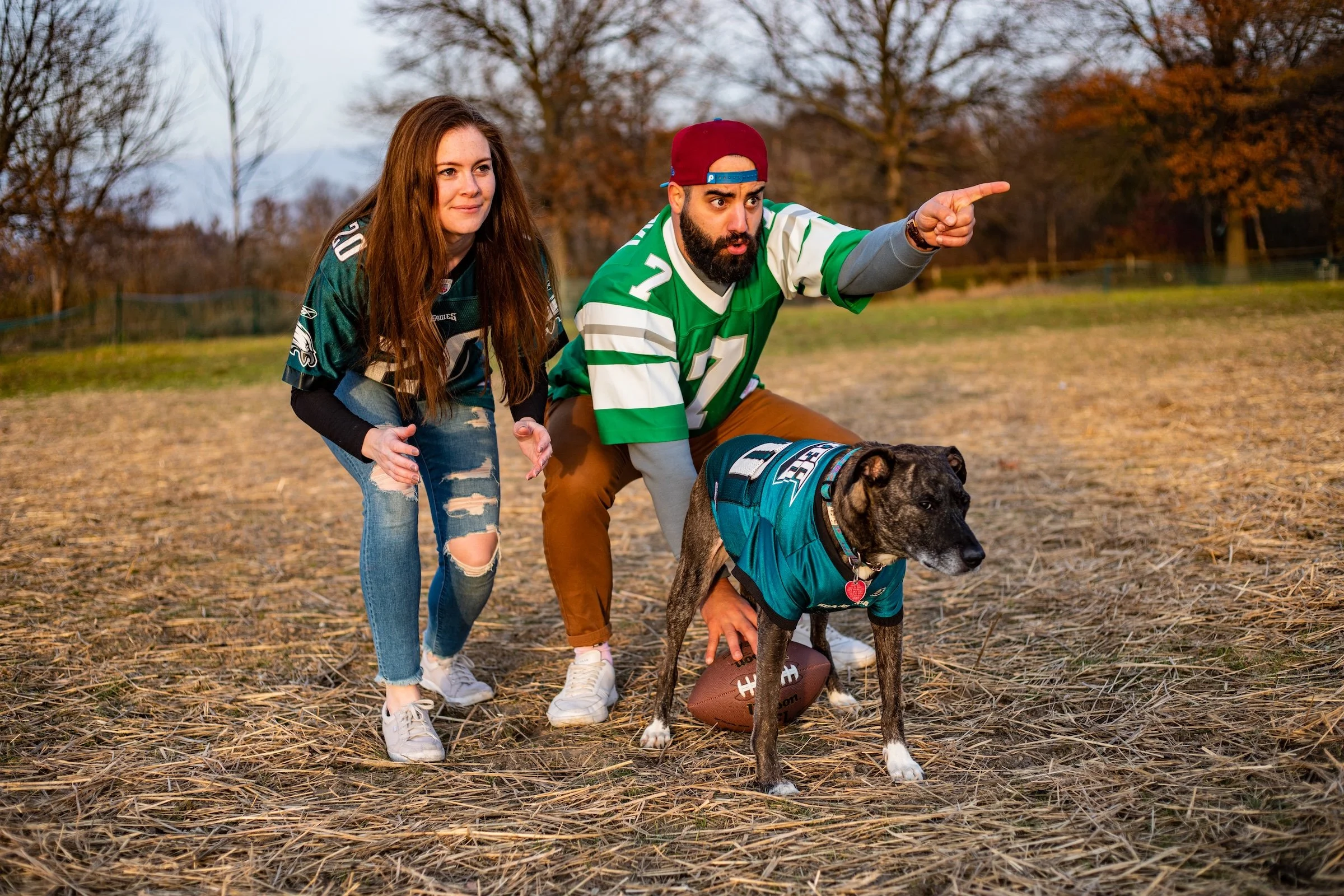 A man, woman, and dog on a football field outdoors. The man is wearing a green sports jersey, a backwards baseball cap, and brown pants, and appears to be coaching or giving directions. The woman is wearing a Philadelphia Eagles jersey and ripped jea