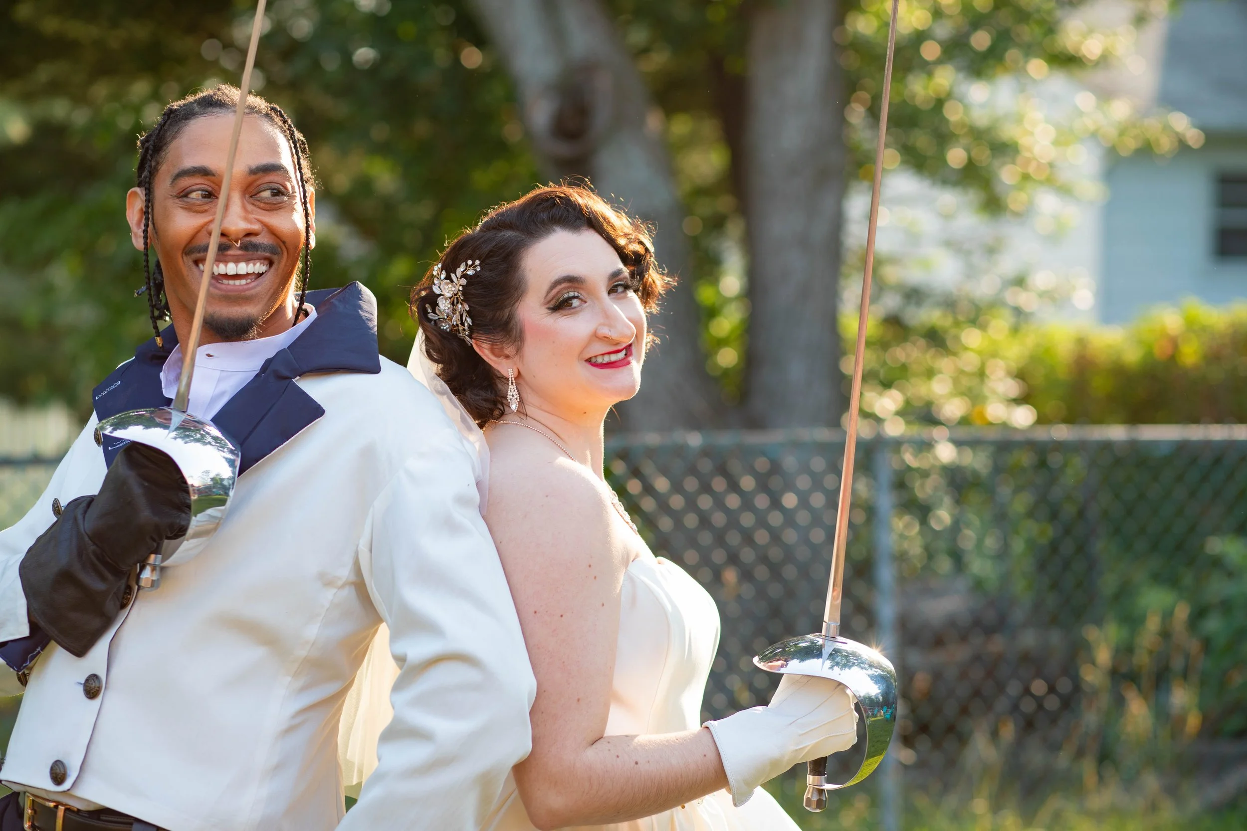 A recently married couple dressed in fencing gear holding swords, smiling outdoors with trees and a fence in the background.