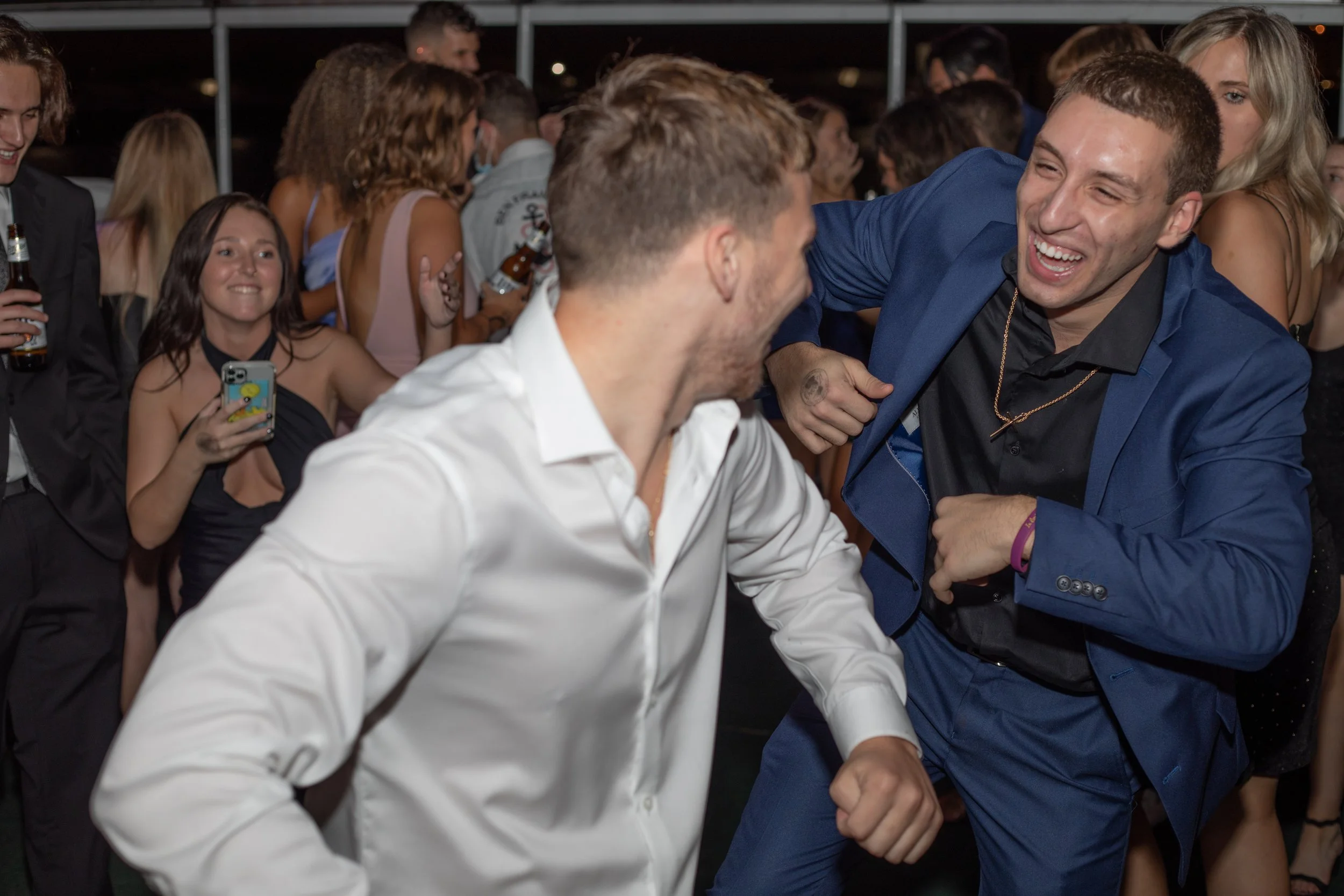 Two men dancing and laughing at a party with a crowd of people in the background.