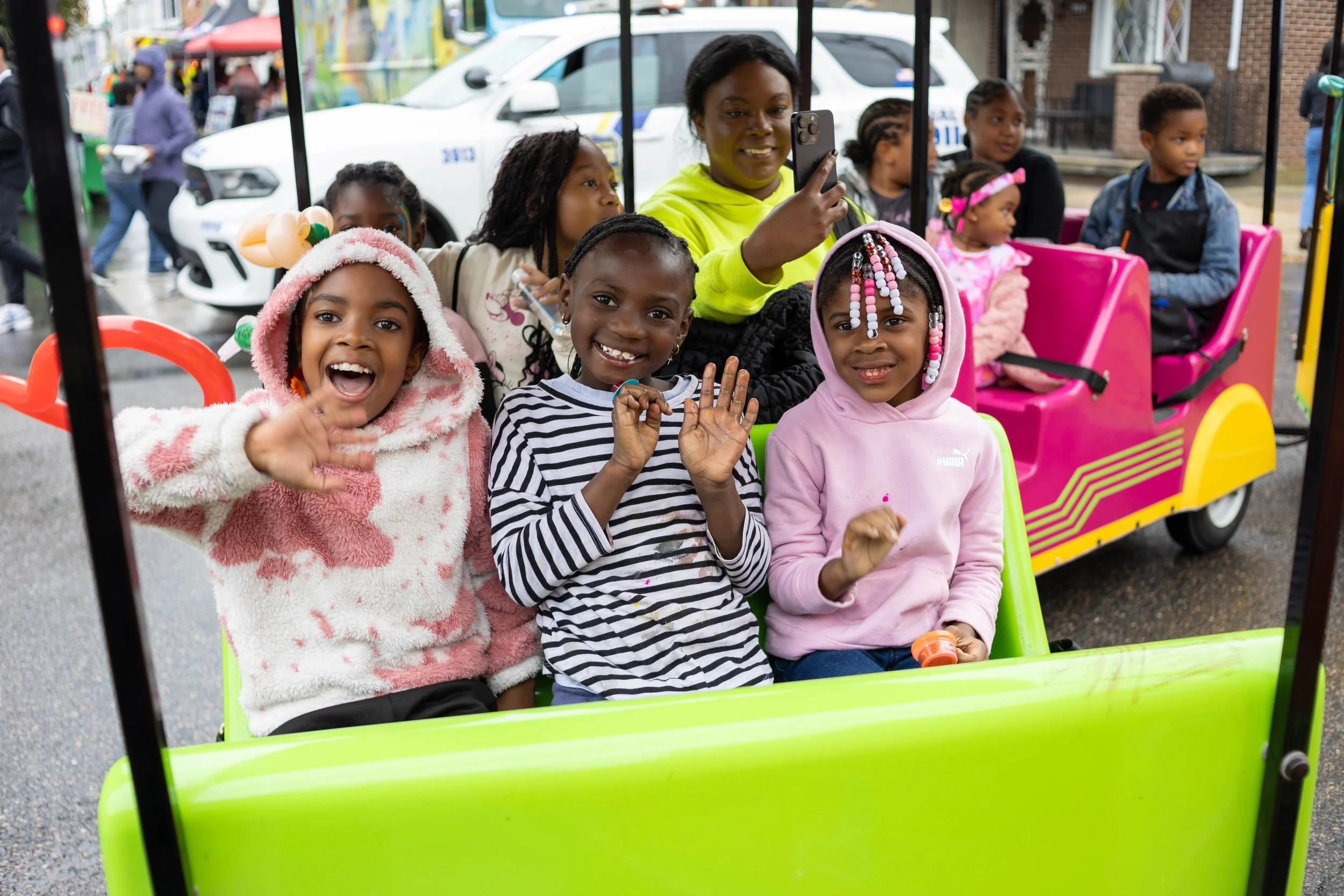 Children sitting in a colorful tram, smiling and waving at the camera, on a street with parked cars and vendor stalls in the background.