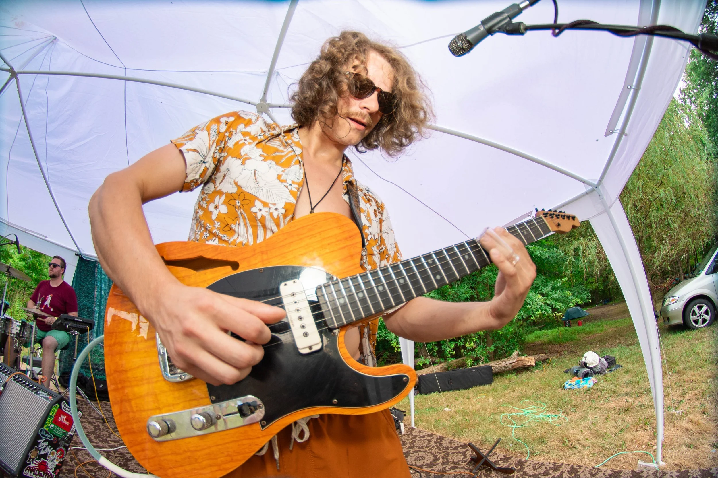 A man with curly hair, sunglasses, and a floral shirt playing an electric guitar inside a white canopy outdoors, with a drummer in the background.