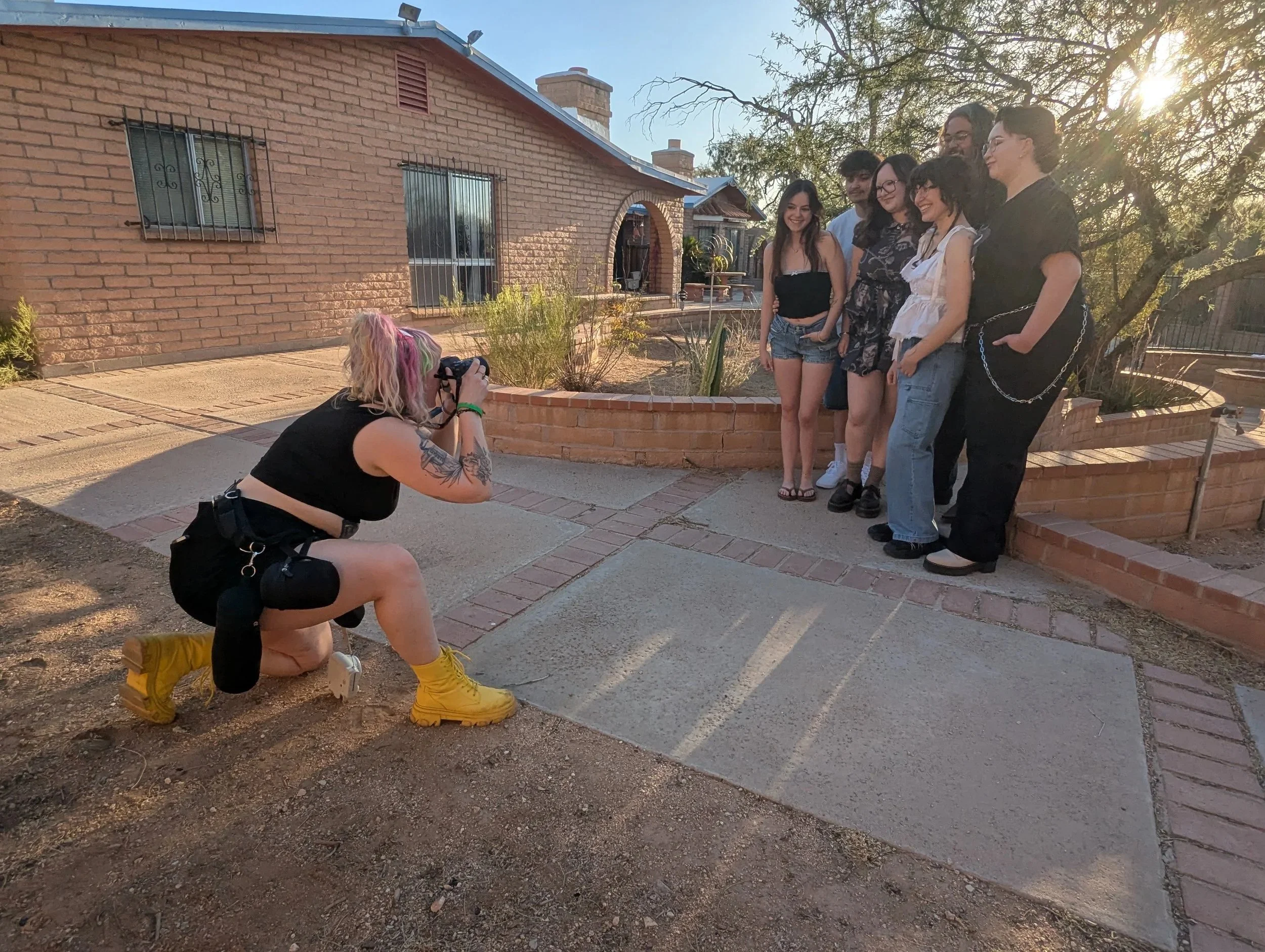 A group of seven young people posing for a photo outdoors on a sunny day. A photographer kneels in front, taking their picture, with a brick house and a tree in the background.