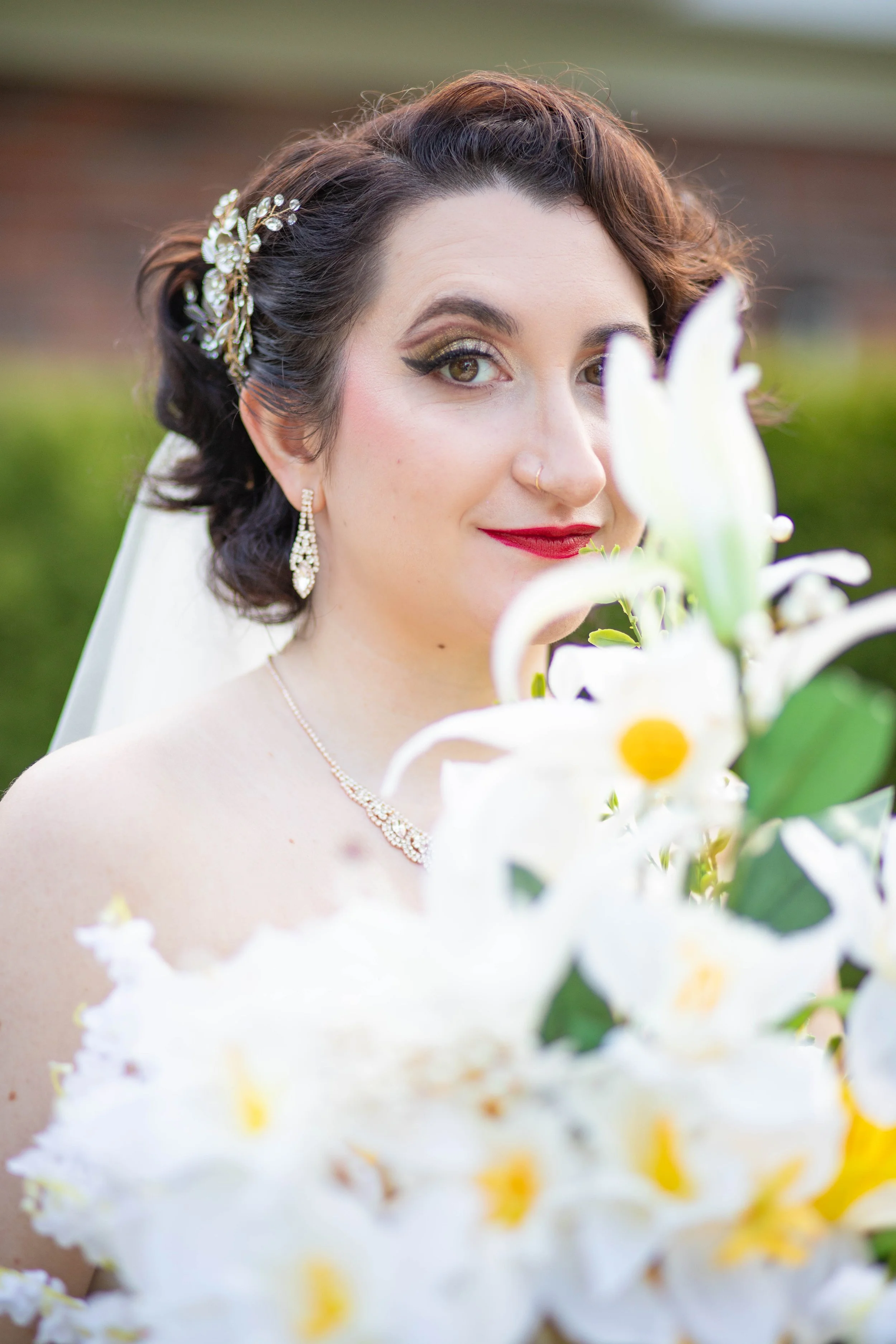Close-up of a woman with dark wavy hair, wearing wedding jewelry, holding a bouquet of white lilies, smiling at the camera outdoors.