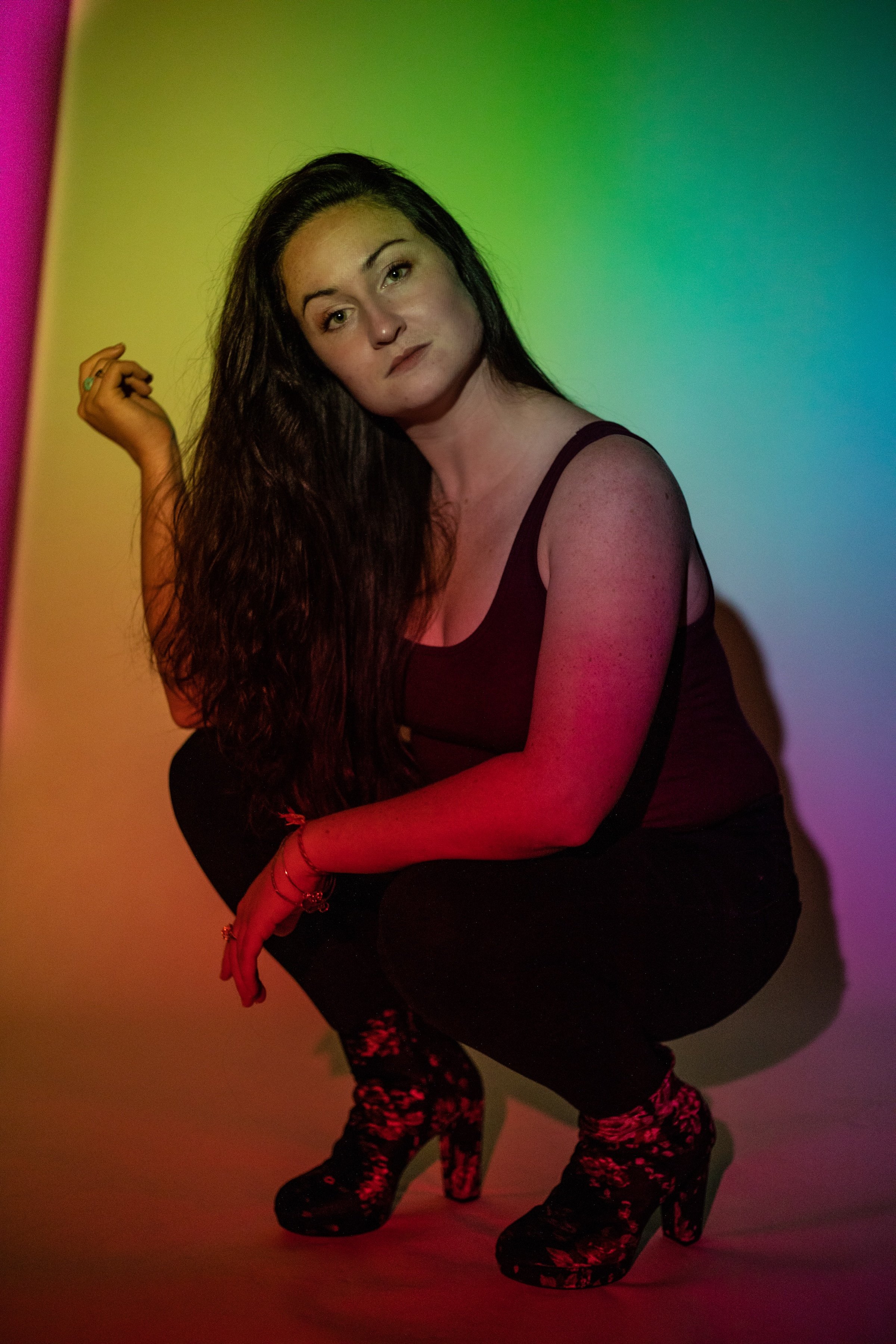 A woman with long dark hair, wearing a black tank top and black pants, squatting in front of a colorful background with rainbow lighting.