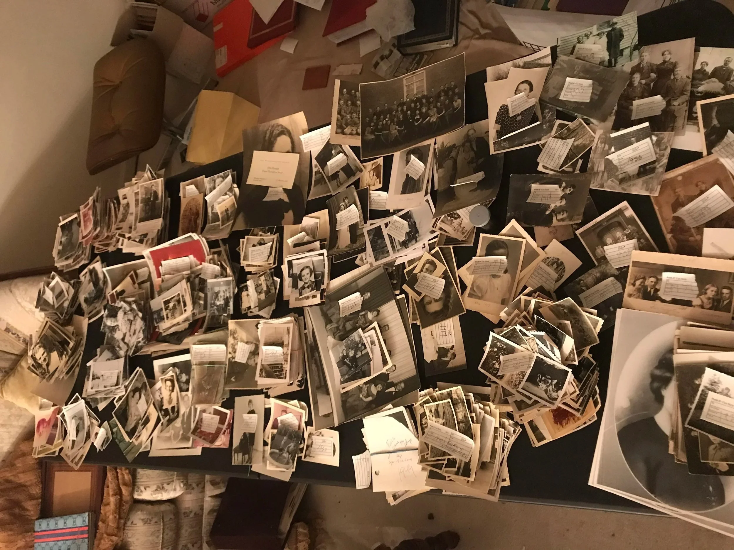 A table cluttered with numerous black-and-white and color photographs, along with some loose papers and notes, in a dimly lit room.