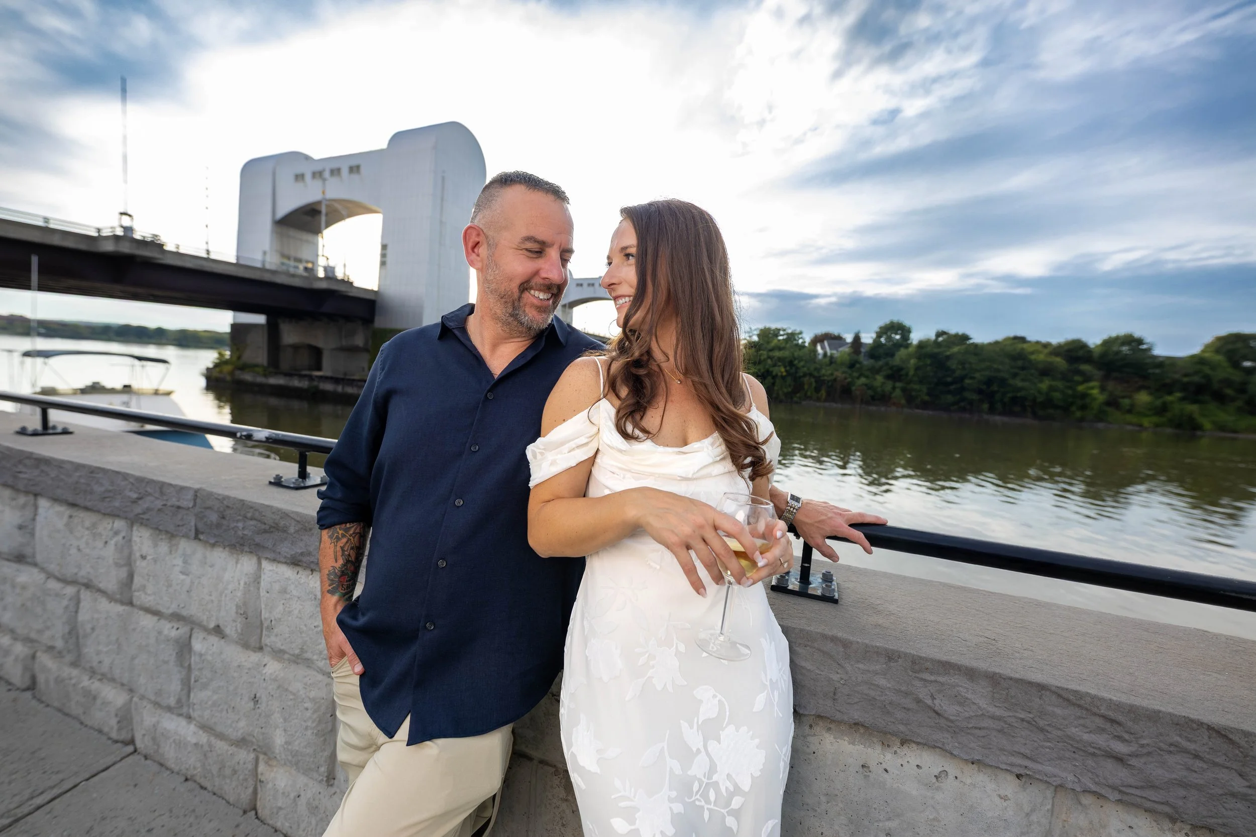 A couple standing on a riverside walkway, smiling and looking at each other, with a bridge and river in the background during the evening.