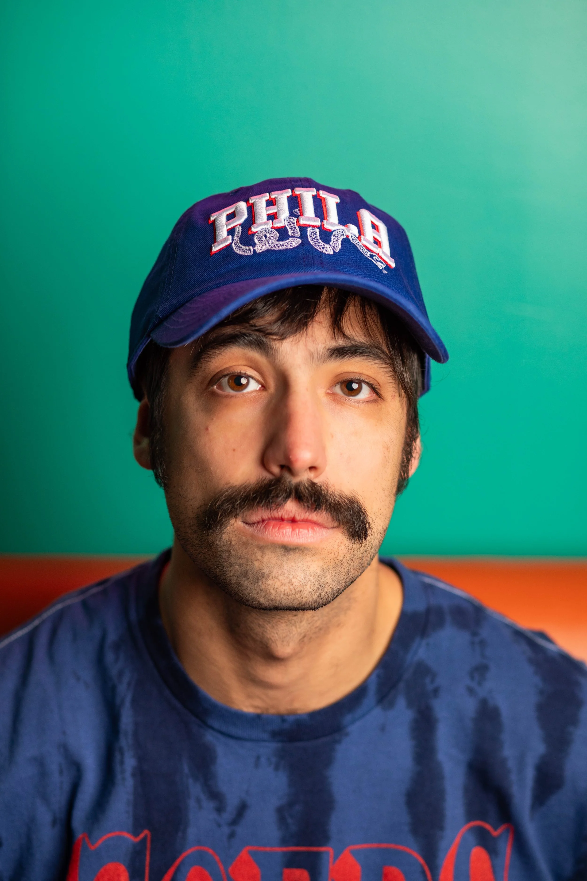 A young man with dark hair, brown eyes, and a thick mustache, wearing a blue Chicago Cubs cap and a tie-dye blue t-shirt, sitting against a green and orange background.