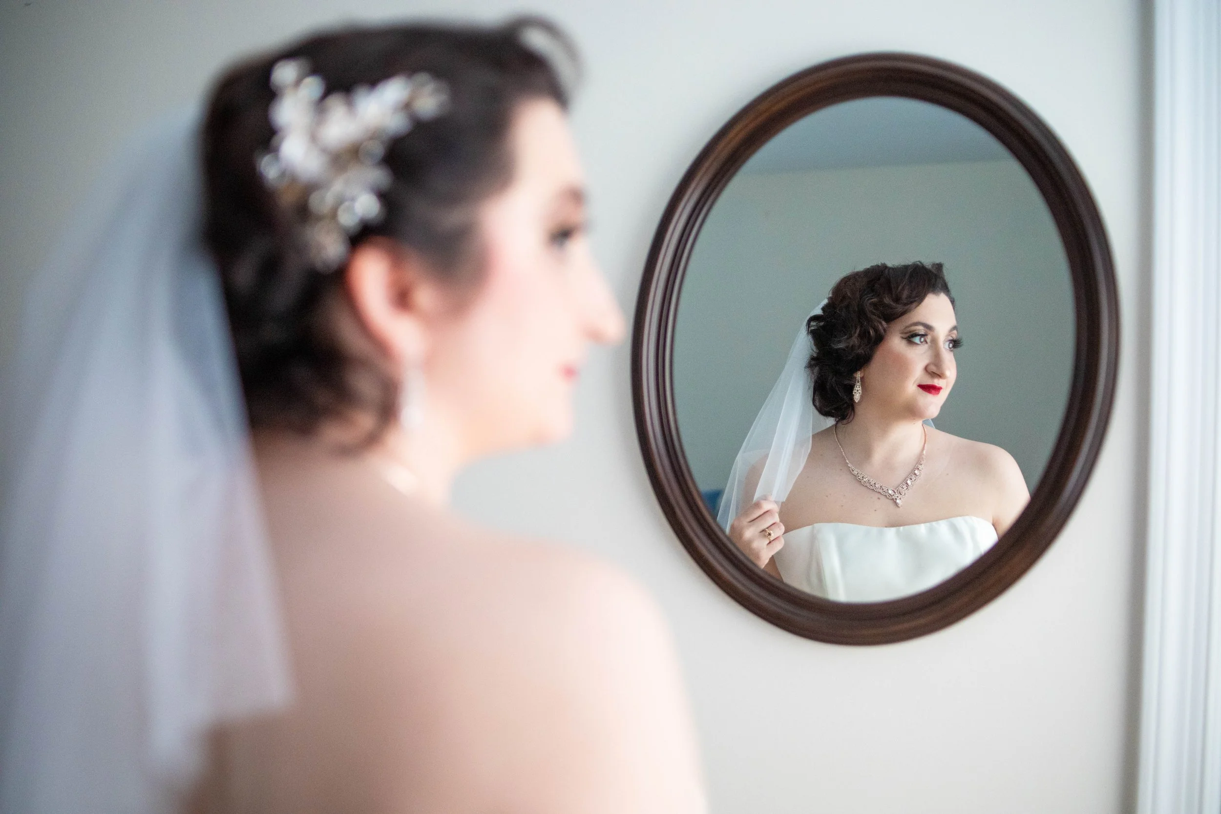 A bride looking at herself in a mirror, wearing a strapless wedding dress, veil, and jewelry, with her reflection visible in a round mirror.