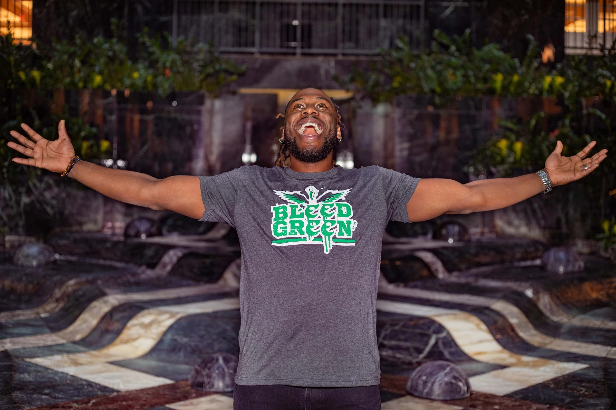 A joyful man with outstretched arms standing in front of a decorative water fountain with greenery, smiling broadly and wearing a gray T-shirt that says 'Bleed Green' with a graphic of a bird.