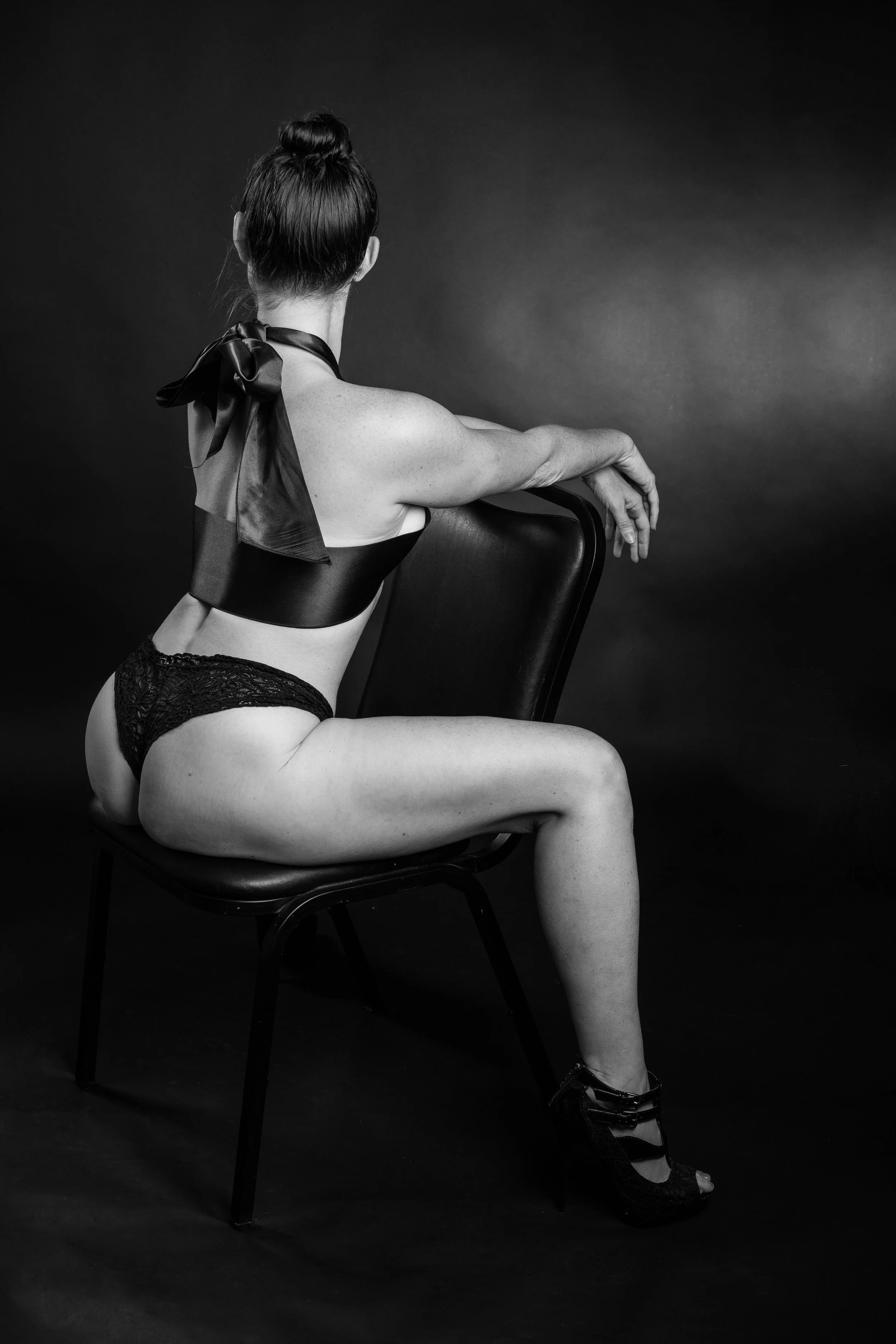 A woman sitting sideways on a chair with her back facing the camera, wearing lingerie and high heels, with her hair in a bun, in a black-and-white portrait.